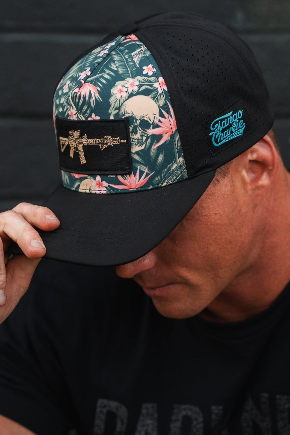 A person dons an Aloha AR Hydro Snapback Hat by Tango Charlie Apparel, featuring a floral and skull-and-rifle design, concealing their face with it as their head tilts slightly downward.