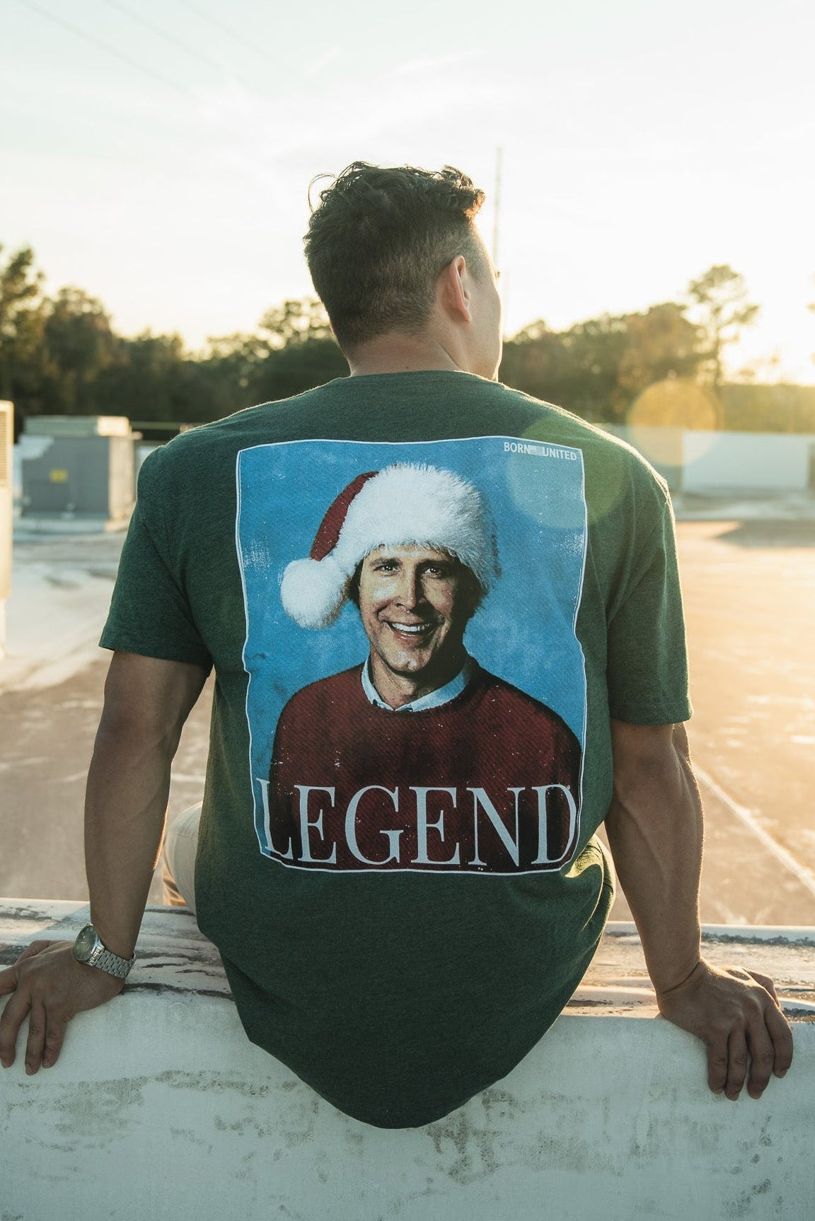 Seated outdoors on a ledge, a man faces away in the sunlit scene framed by trees and clear skies. He sports the Legend - Griswold retro tee by Born United Private Label, featuring a smiling person in a Santa hat labeled LEGEND, capturing the festive spirit like classic Christmas movie moments.