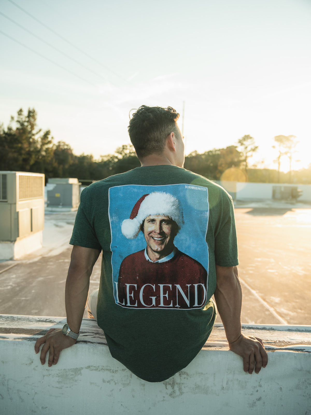 Seated outdoors on a ledge, a man faces away in the sunlit scene framed by trees and clear skies. He sports the Legend - Griswold retro tee by Born United Private Label, featuring a smiling person in a Santa hat labeled LEGEND, capturing the festive spirit like classic Christmas movie moments.
