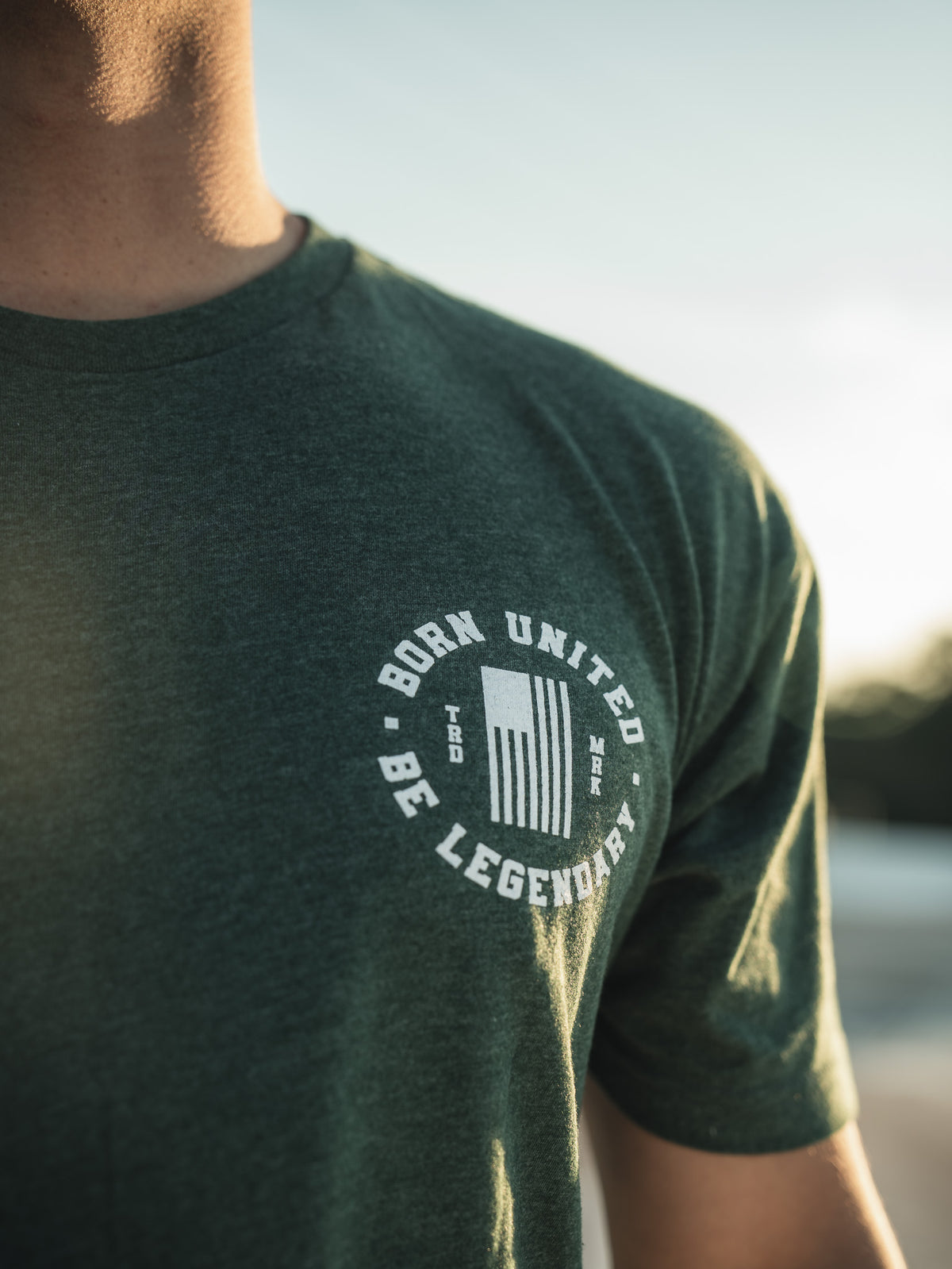 Wearing a Legend - Griswold tee by Born United Private Label, featuring white text BORN UNITED BE LEGENDARY, a person stands against a blurred backdrop of greenery and sky, creating an outdoor ambiance.
