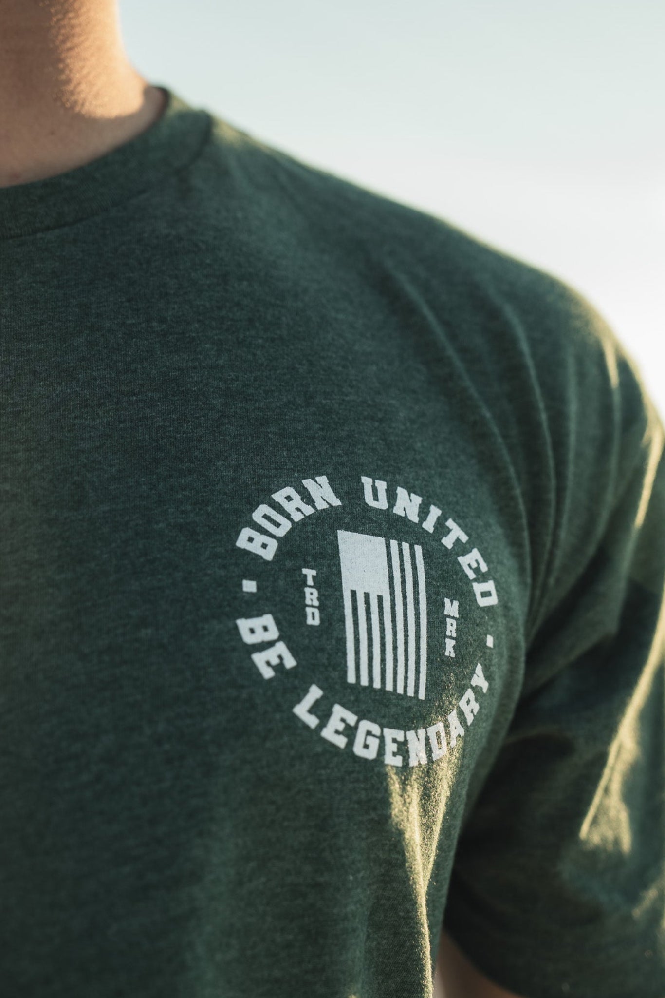 Wearing a Legend - Griswold tee by Born United Private Label, featuring white text BORN UNITED BE LEGENDARY, a person stands against a blurred backdrop of greenery and sky, creating an outdoor ambiance.