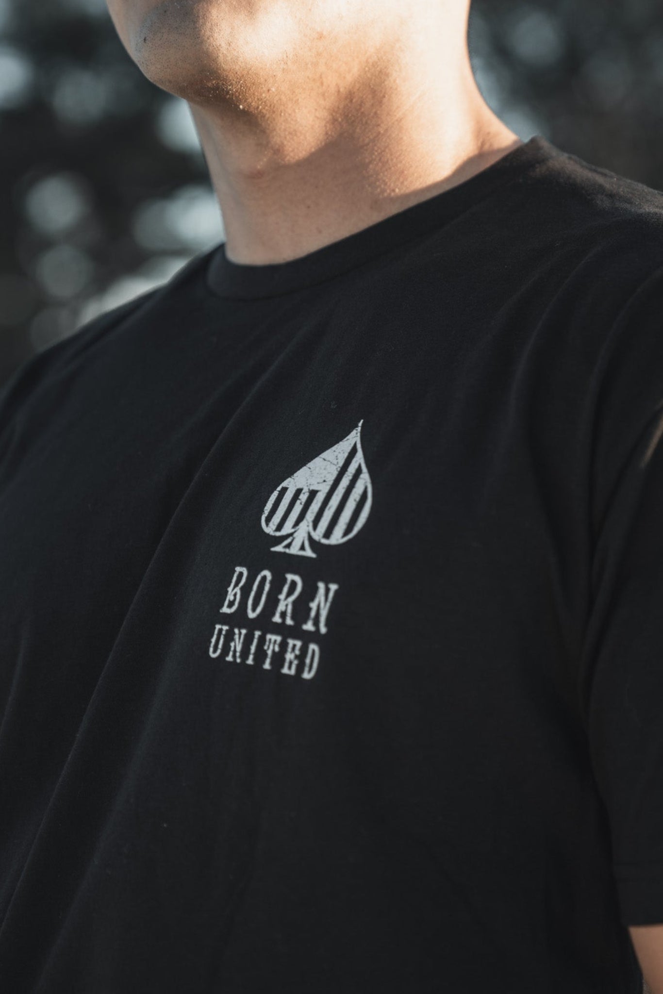 A person wearing a black Angel of Death T-shirt by Born United Private Label stands outdoors, with trees and a clear sky blurring behind them. The shirt features an upward-pointing spade and BORN UNITED. Only the torso and part of the face are visible, evoking the presence of an AC-130 aircraft above.