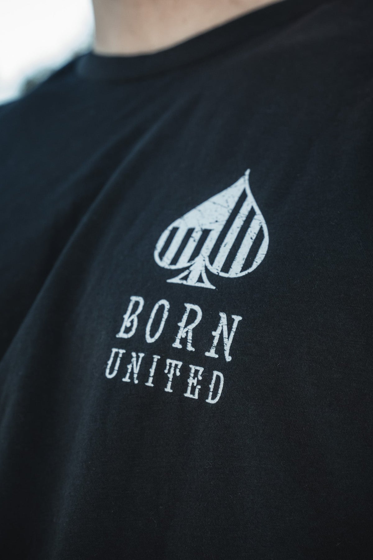 A person wears a Born United Private Label Angel of Death black T-shirt, featuring an abstract spade-like design reminiscent of an AC-130. Vertical stripes are above the distressed font words BORN UNITED.