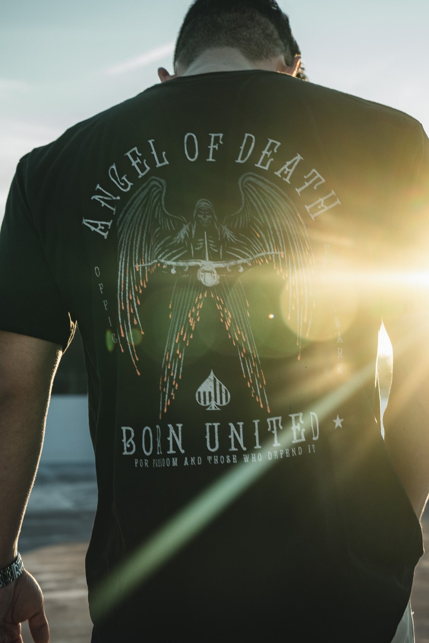 A person wearing Born United Private Labels Angel of Death black T-shirt, featuring an AC-130 military aircraft graphic under a fiery sky, stands with their back to the camera. The sunset lens flare on the right illuminates the design.