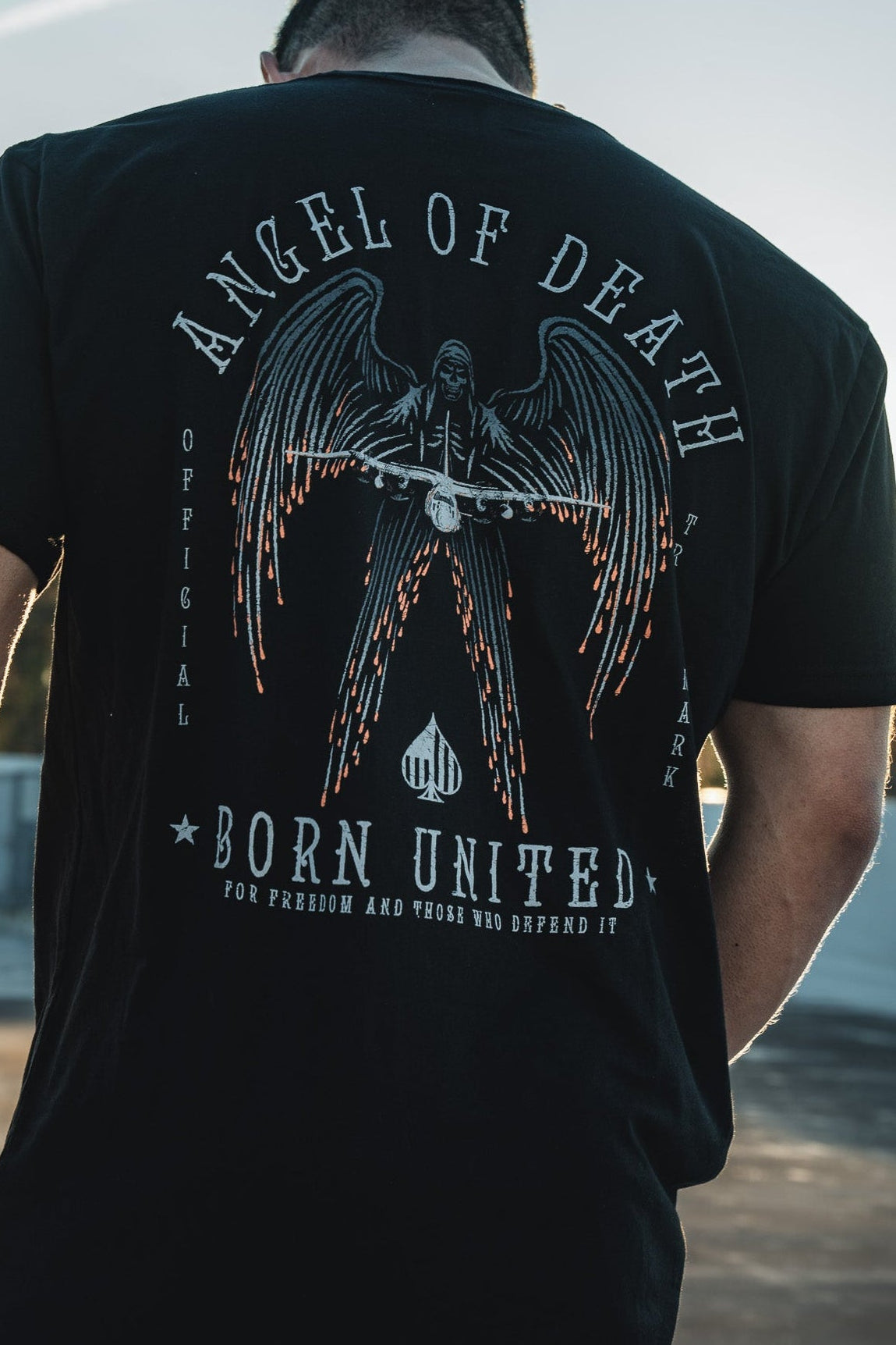 A man faces away from the camera, wearing a black Angel of Death T-shirt by Born United Private Label. The graphic shows an angel with swords, evoking the legendary AC-130 aircraft. Hes outdoors under a wide blue sky.