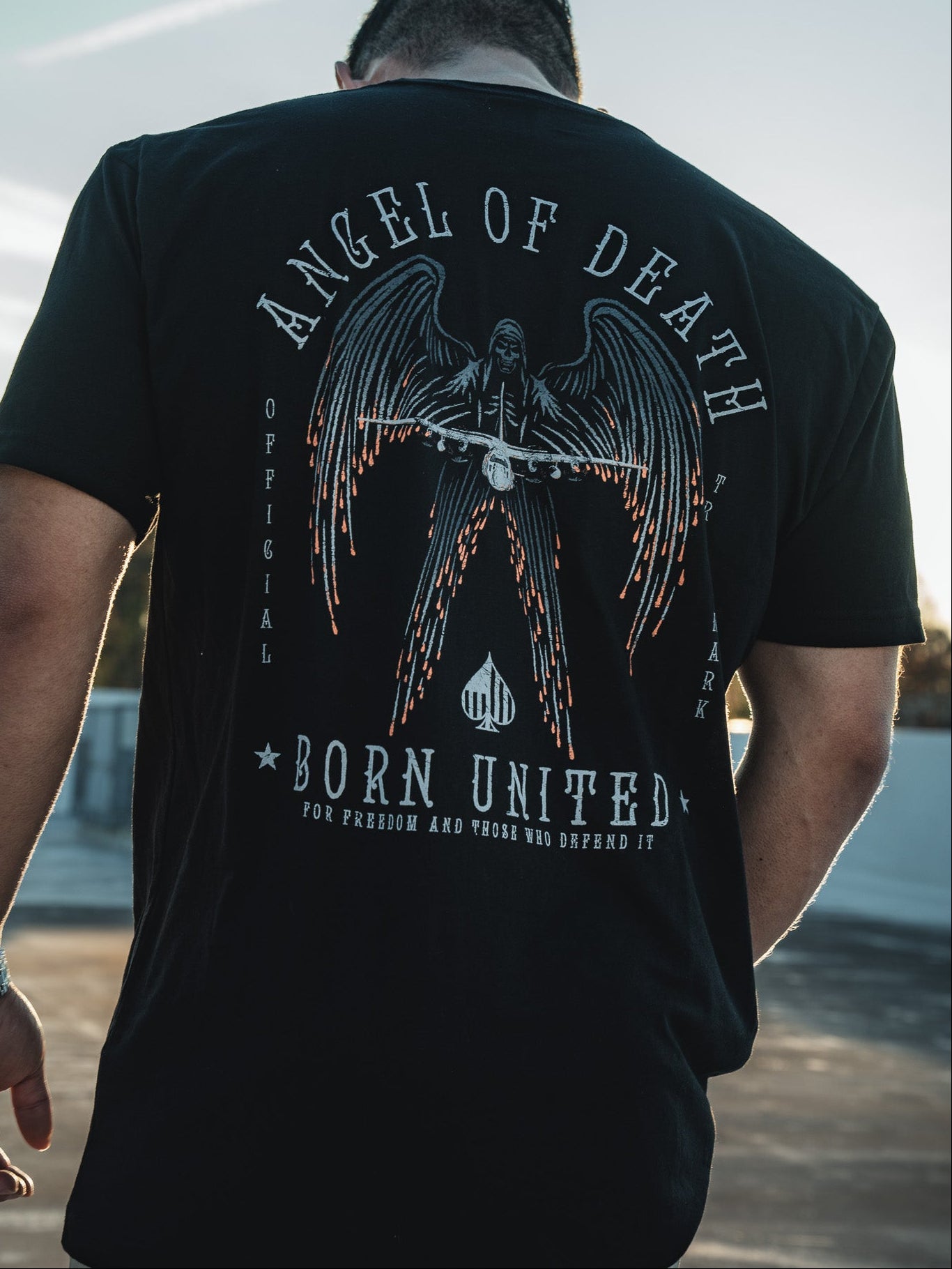 A man faces away from the camera, wearing a black Angel of Death T-shirt by Born United Private Label. The graphic shows an angel with swords, evoking the legendary AC-130 aircraft. Hes outdoors under a wide blue sky.