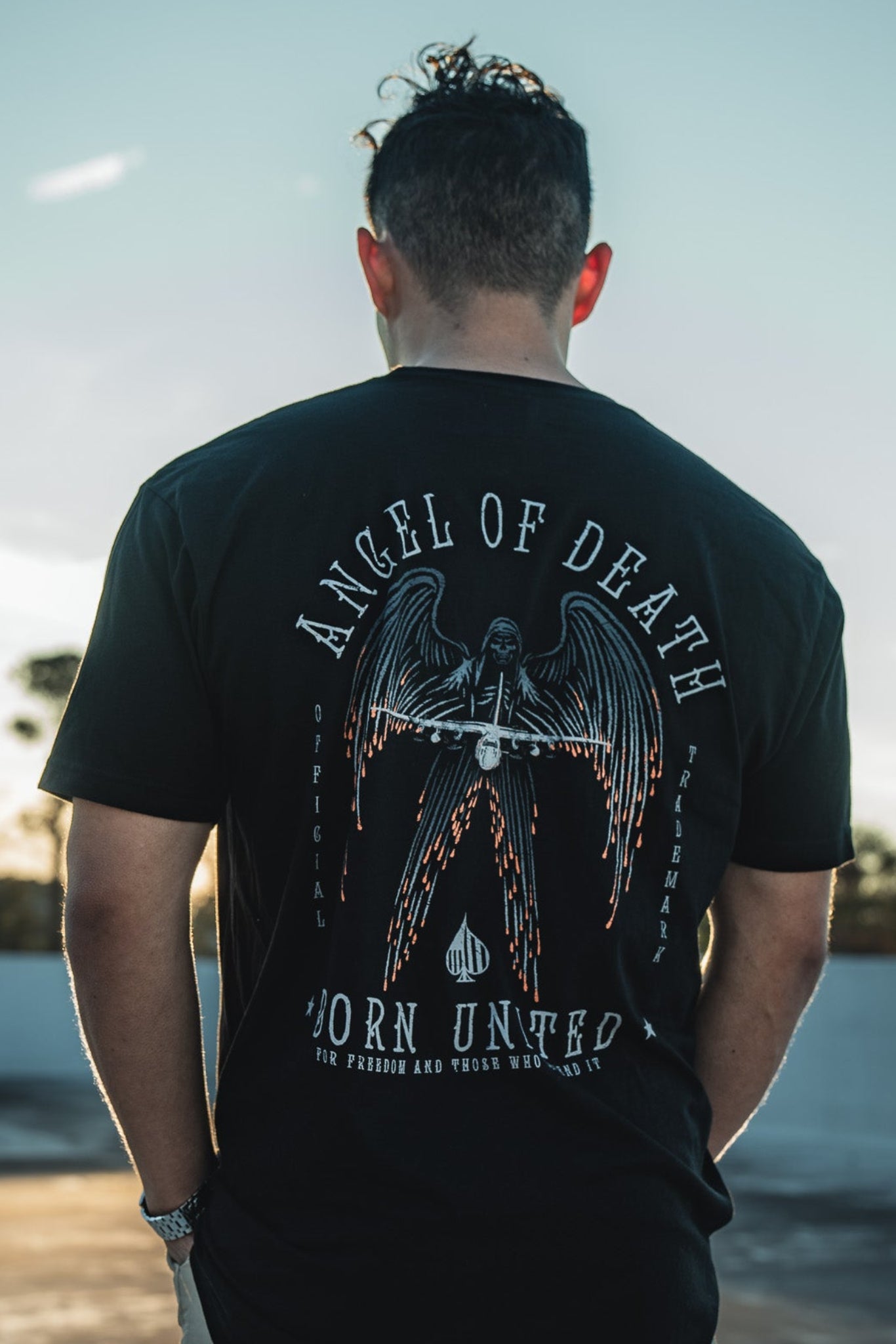 A man with curly hair stands on a rooftop, facing away and wearing a Born United Private Label black T-shirt featuring the Angel of Death design. With a clear sky and scattered clouds, it creates a serene backdrop for this striking image.