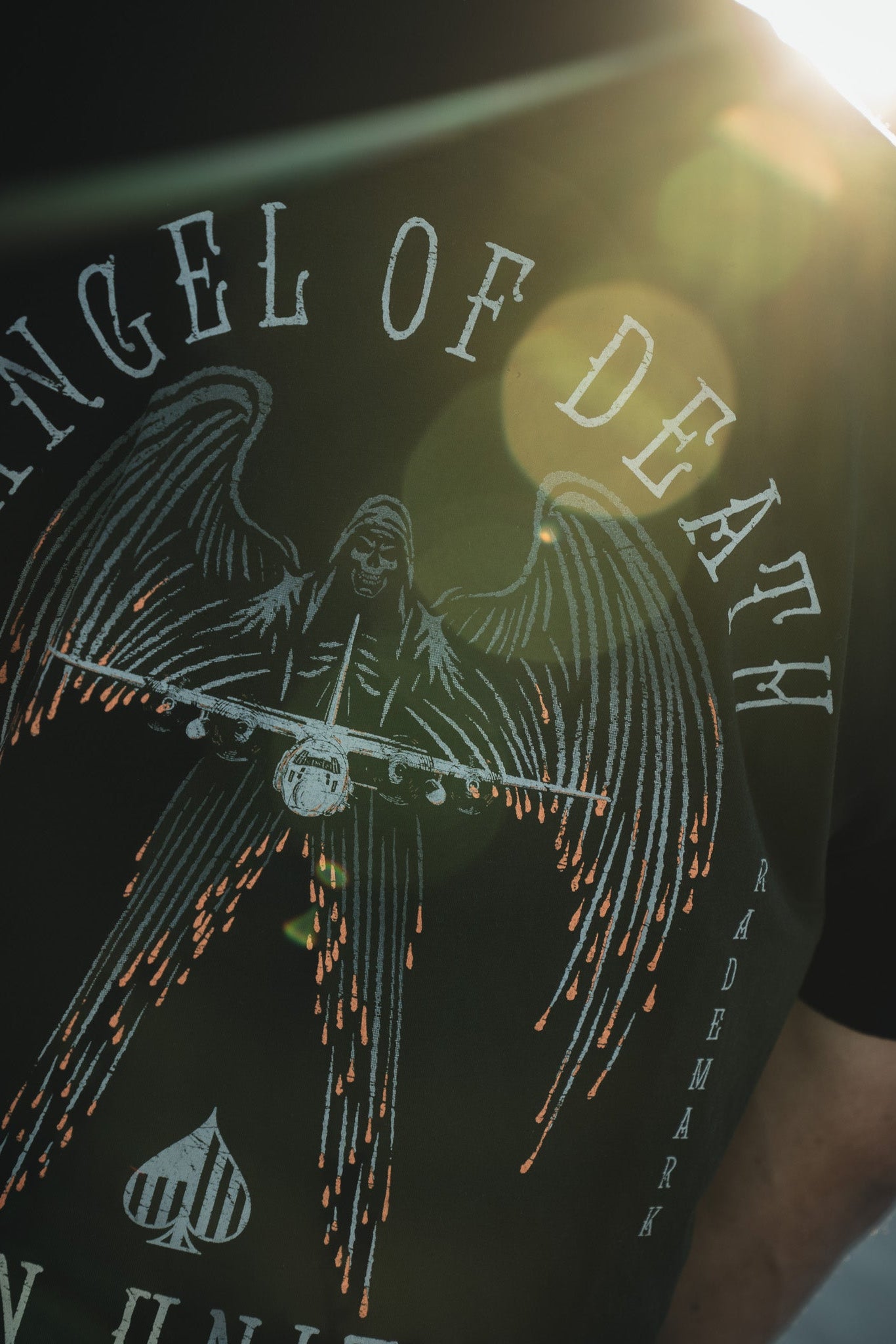 A person in a Born United Private Label black shirt displays the Angel of Death design, featuring an angel with large wings holding an AC-130 aircraft. The text ANGEL OF DEATH, TRADEMARK appears, with a backlit lens flare adding drama.
