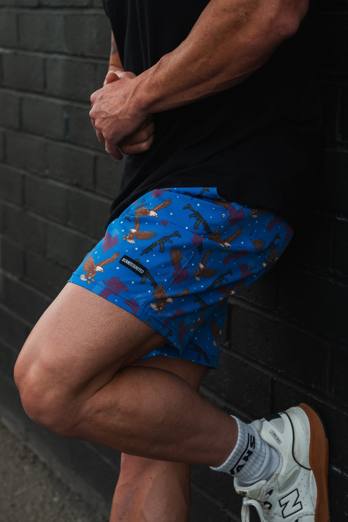 A person leans against a dark brick wall, displaying the lower half of their outfit. They wear a black shirt and colorful blue Mens Performance Hybrid Shorts | 5.5- Merica Baby with bird patterns from Born United Private Label, perfect for gym-to-swim activities, paired with white socks and New Balance sneakers.