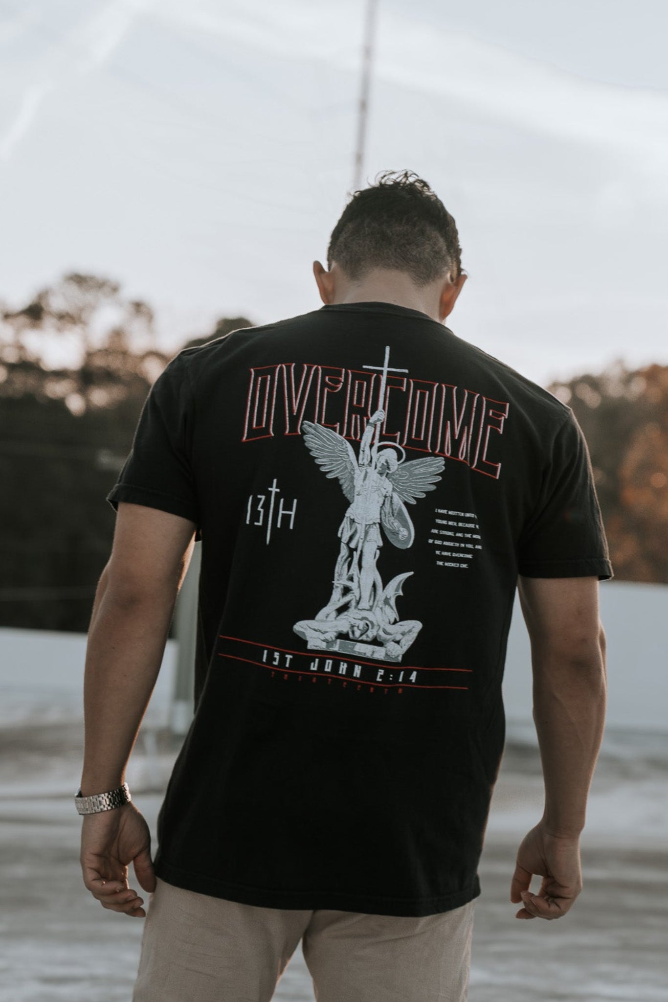A young man stands outdoors with his back to the camera, wearing the Overcome Premium Tee by 13th. The black t-shirt features an angel graphic and Word of God, set against a serene backdrop of sunlit trees and light-colored ground.