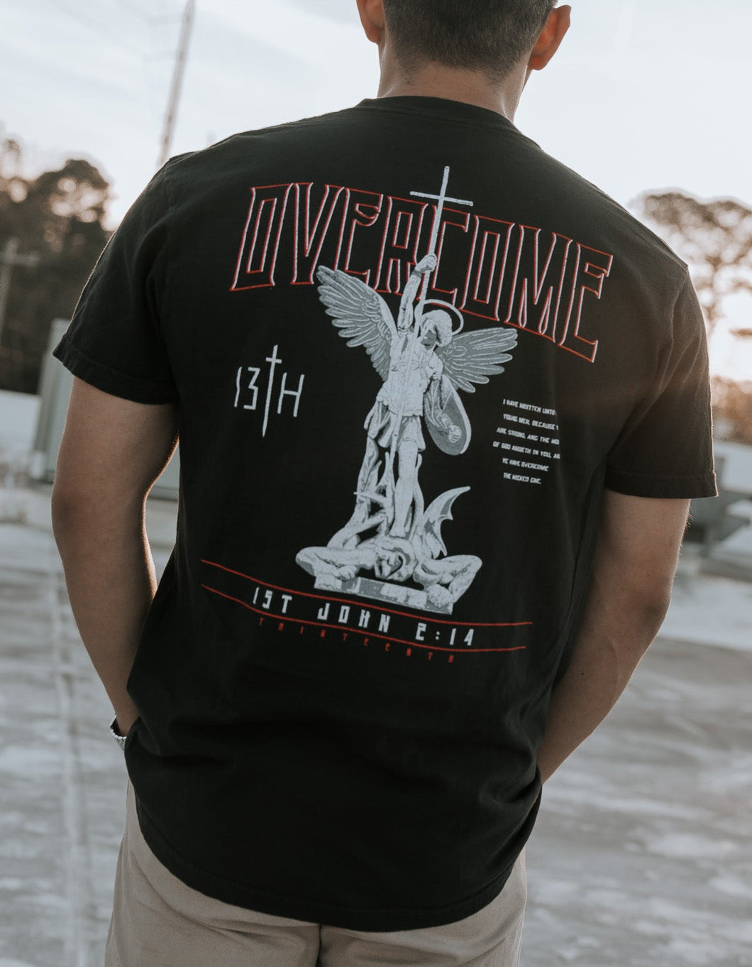 On a rooftop against a clear sky with contrails, a young man faces away wearing the Overcome Premium Tee by 13th. The black T-shirt features a large angel graphic and the words Overcome and 1st John 4:17, reflecting the word of God, with trees in the background.