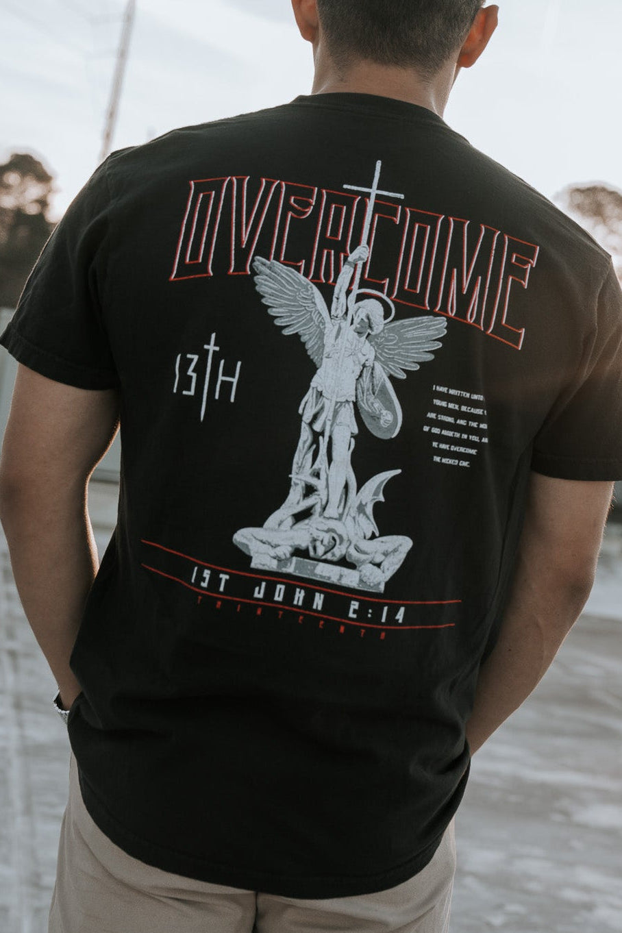 On a rooftop against a clear sky with contrails, a young man faces away wearing the Overcome Premium Tee by 13th. The black T-shirt features a large angel graphic and the words Overcome and 1st John 4:17, reflecting the word of God, with trees in the background.