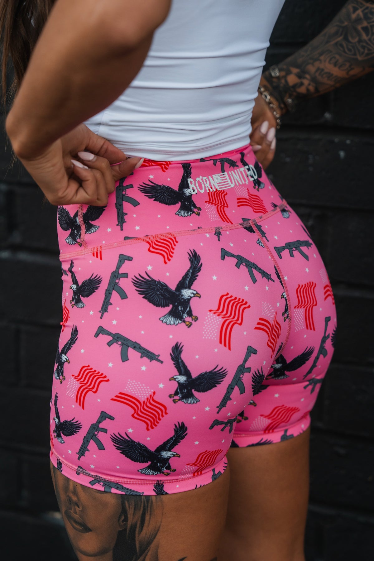 Wearing Born United Private Labels Womens High-Waisted Seamless Front Compression Training Shorts | Pink Victory, adorned with eagles, rifles, and American flags, she pairs them with a white top against a dark wall as tattoos peek from beneath the breathable fabric.