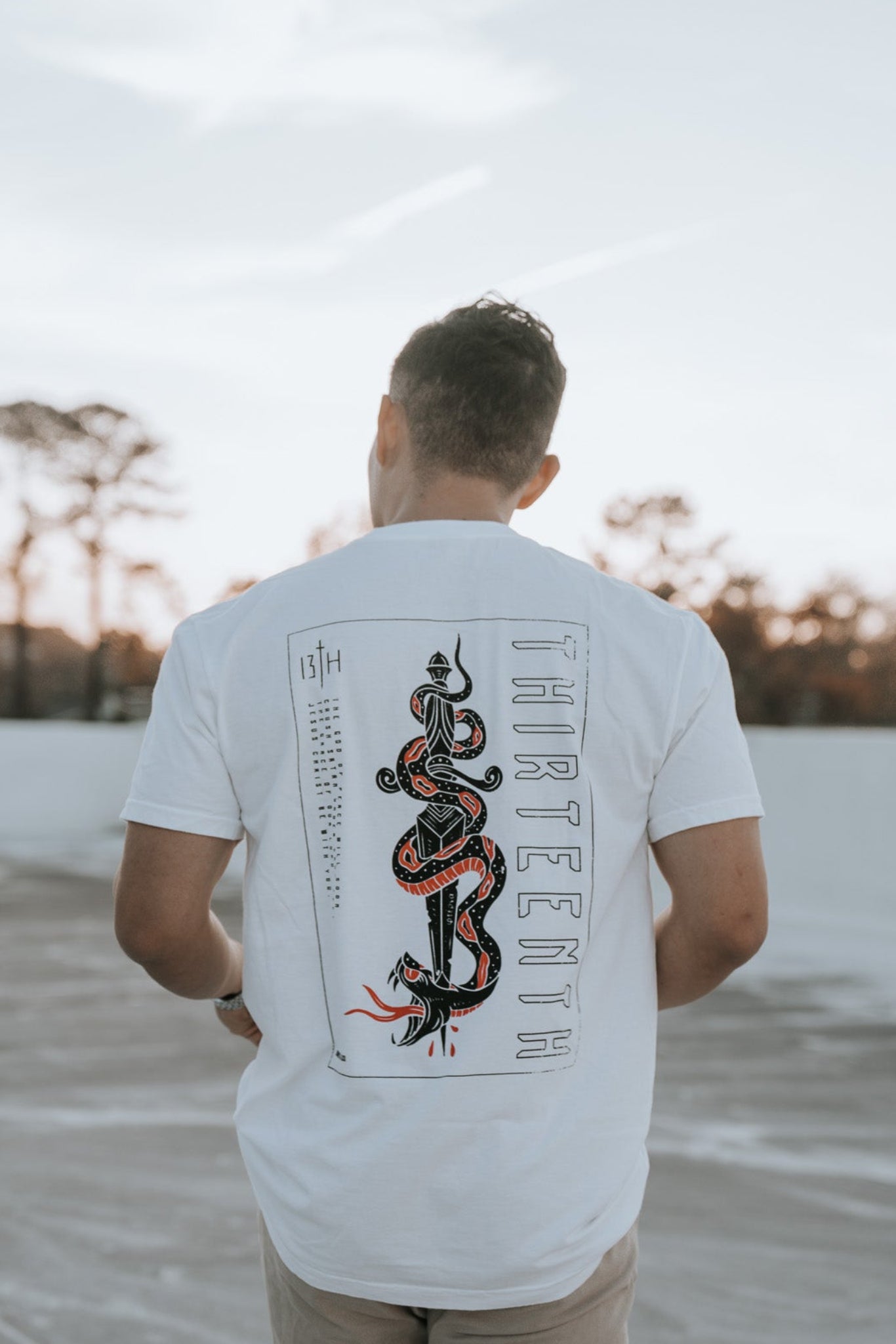 A man with short hair stands on a concrete surface outdoors, facing away. Hes wearing a 13th brand Lucifers Demise Premium Tee in white featuring a snake graphic and the words Crush Satan on the back. The sky is clear with trees in the serene background.