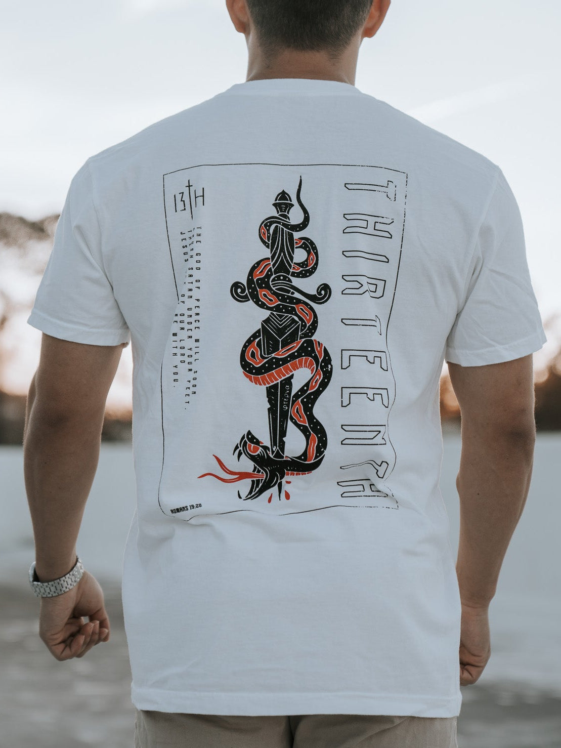 A person with short hair stands outside, wearing the 13th brand Lucifers Demise Premium Tee in white, featuring a red and black snake and THREEFOUR on the back. The clear sky and gently swaying distant trees create a serene scene.