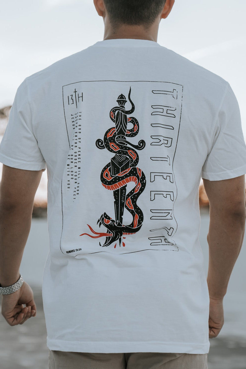 A person with short hair stands outside, wearing the 13th brand Lucifers Demise Premium Tee in white, featuring a red and black snake and THREEFOUR on the back. The clear sky and gently swaying distant trees create a serene scene.