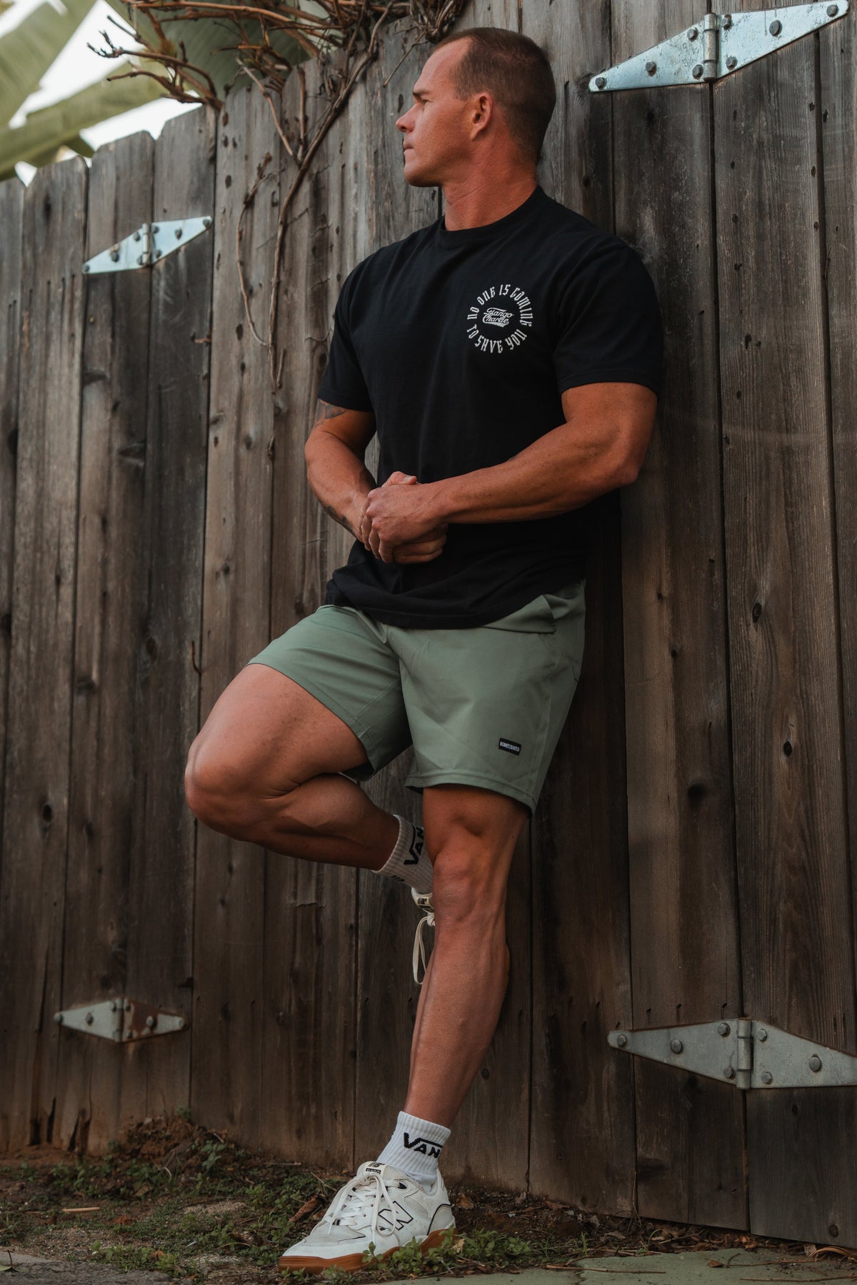 A man leans on a wooden fence, glancing right. Hes wearing a black T-shirt and Mens Standard Performance Shorts | 5.5- OD Green by Born United Private Label, crafted from breathable stretch material. White socks and sneakers complete his relaxed style as plants peek above the metal-hinged fence.