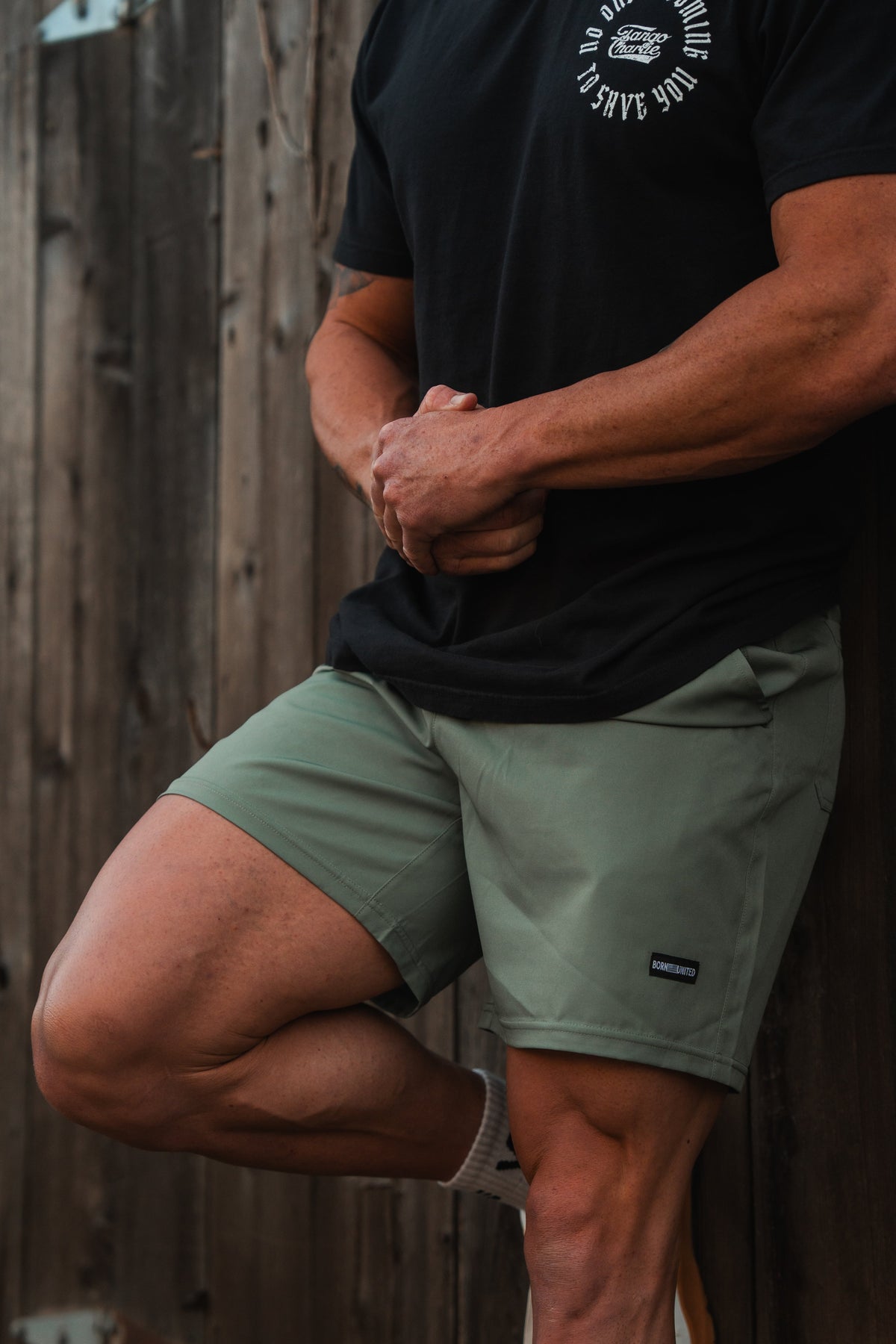 A person in a black t-shirt and Born United Private Labels Mens Standard Performance Shorts | 5.5- OD Green leans against a wooden fence. Their arms are crossed, and they wear white socks. The t-shirt is breathable and stretchable with white text and a chest graphic.