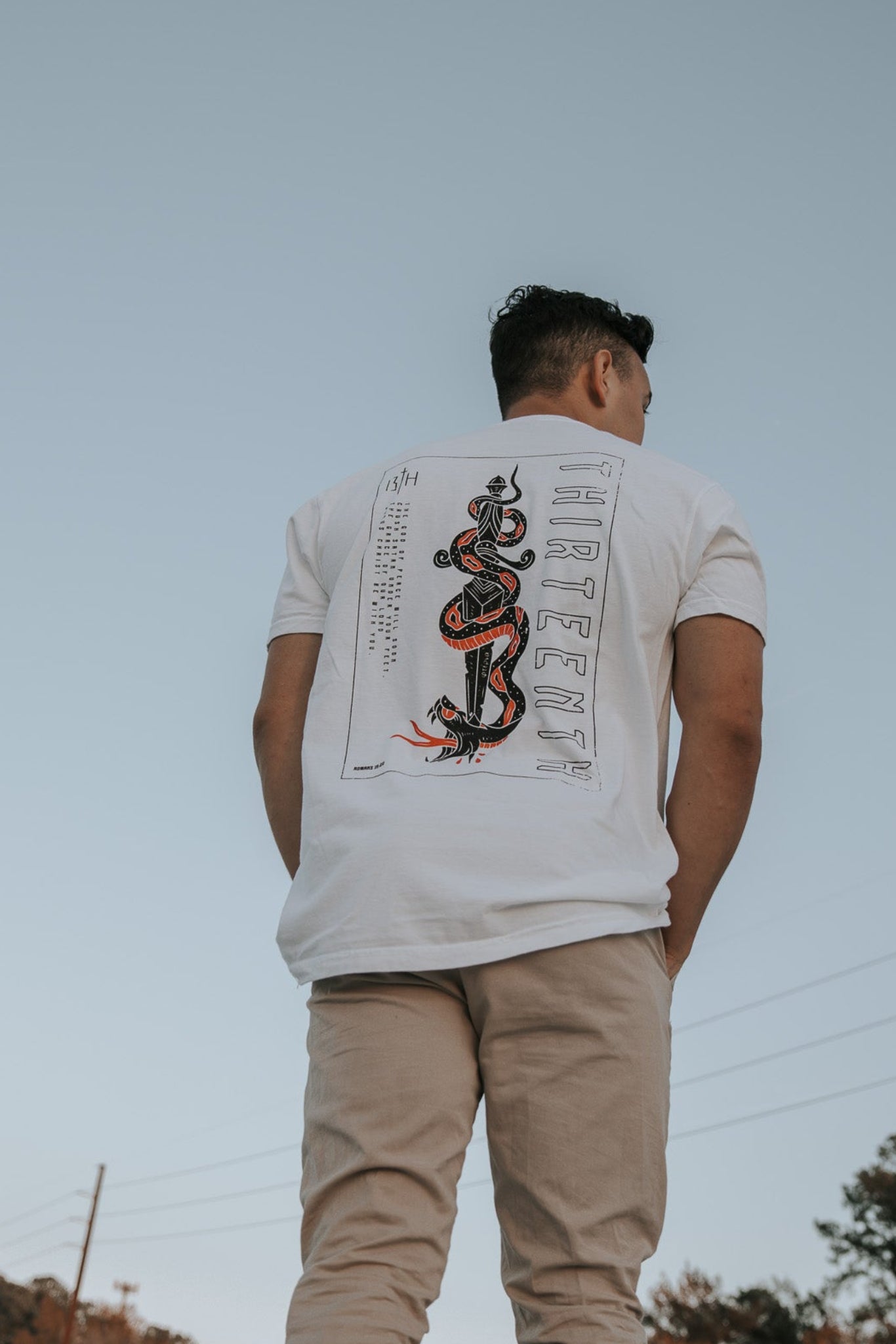 A man wearing the 13ths Lucifers Demise Premium Tee- White with a snake and Crush Satan graphic stands outdoors in beige pants, set against a clear sky with trees and power lines providing a serene background.