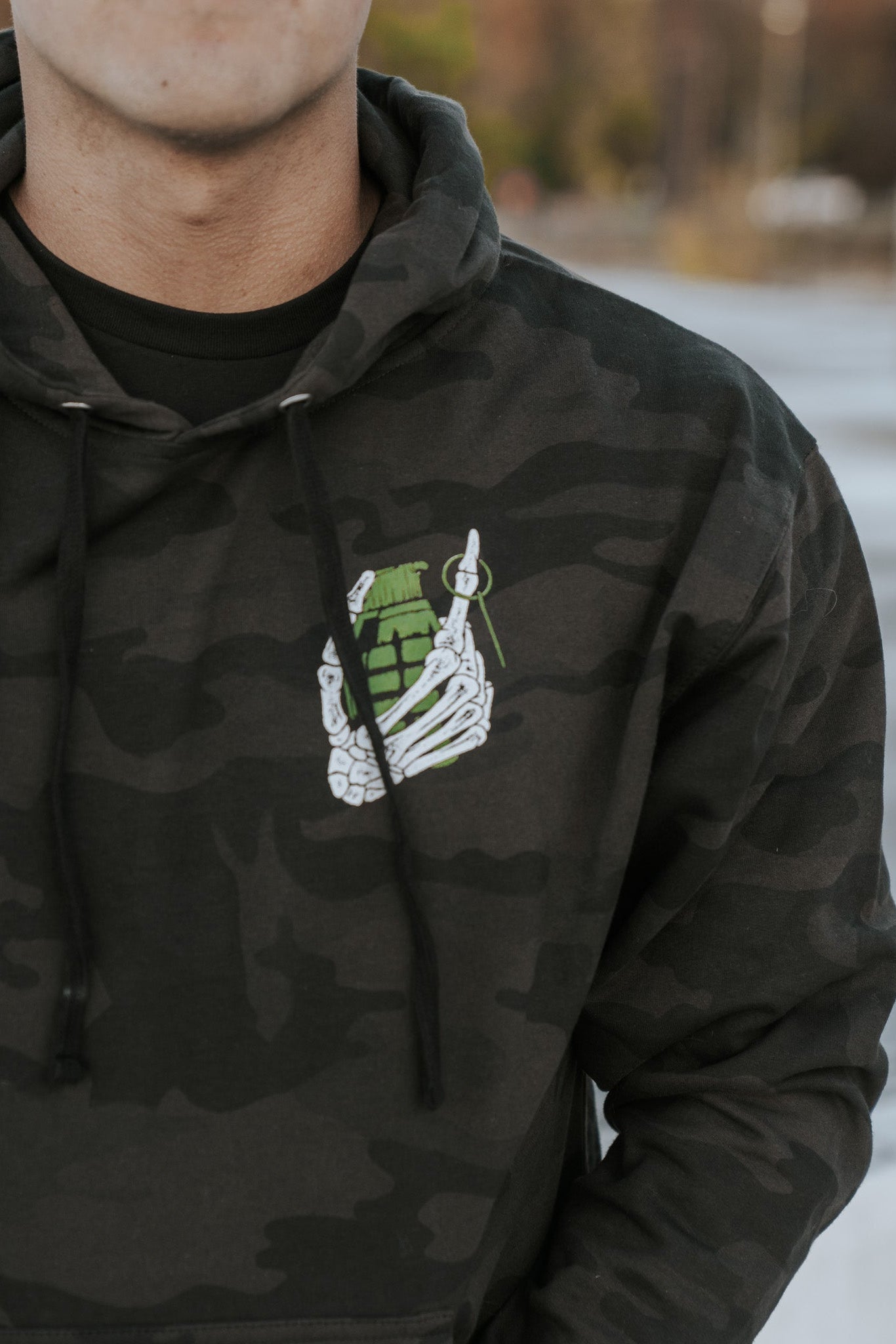 Wearing the Born United Private Labels Full Send Camo Hoodie (Warehouse), featuring a skeletal hand holding a green grenade, the person exudes the Full Send vibe with their face hidden against a blurred outdoor background.
