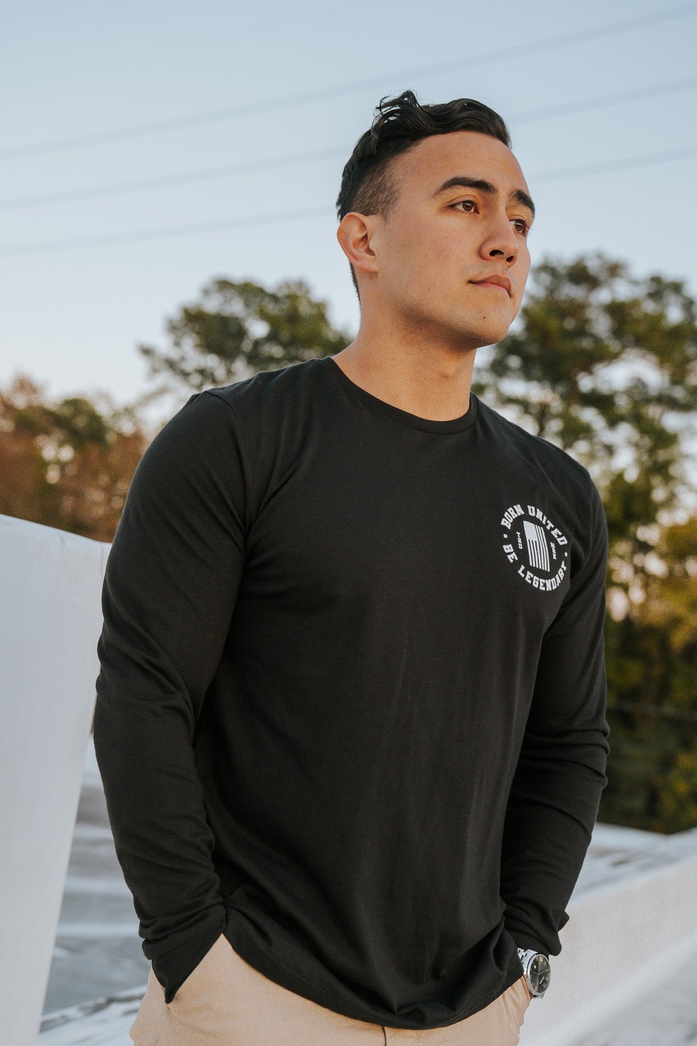 A person stands outdoors on a bridge, donning the Forever Legend Trump - Long Sleeve by Born United Private Label with beige pants, hands in pockets. The picturesque background of trees and a sunset sky enhances the serene atmosphere.