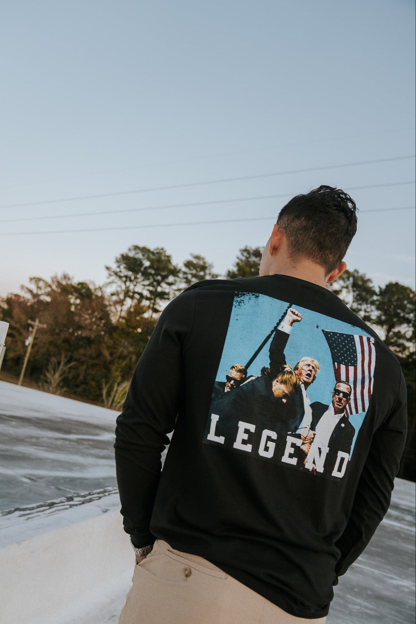 A person outdoors wears a Born United Private Label Forever Legend Trump long-sleeve shirt featuring several people, an American flag, and Forever Legend text. The vivid design is set against trees and a clear sky.