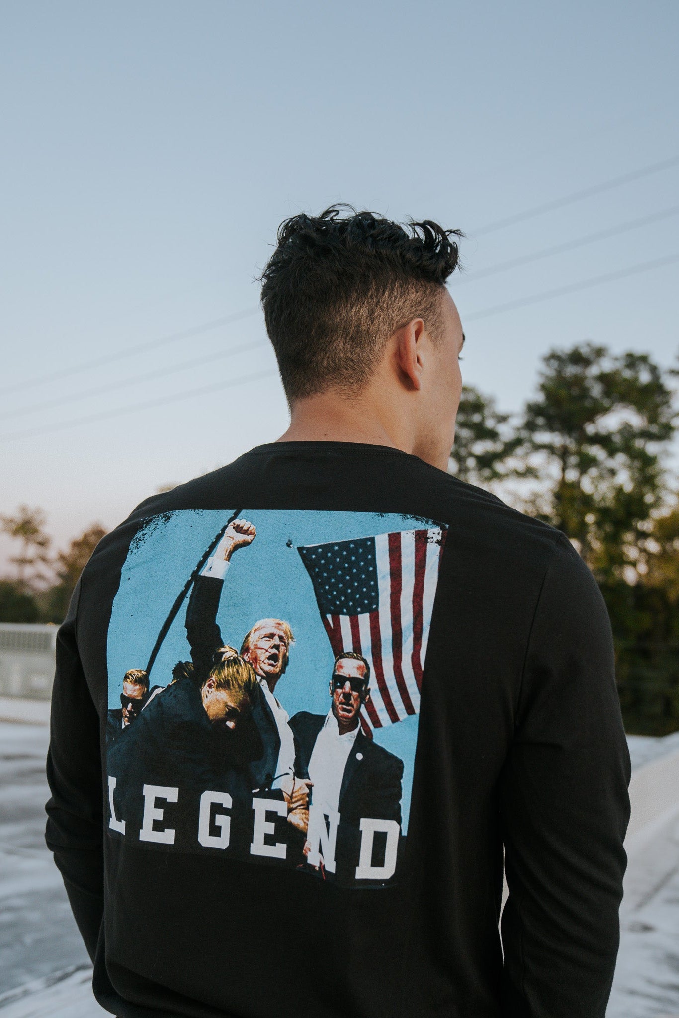 A person with short curly hair, seen from behind, wears the Born United Private Label’s Forever Legend Trump long sleeve featuring people holding an American flag and raising fists. Forever Legend is inscribed below. The background shows trees against a clear sky.