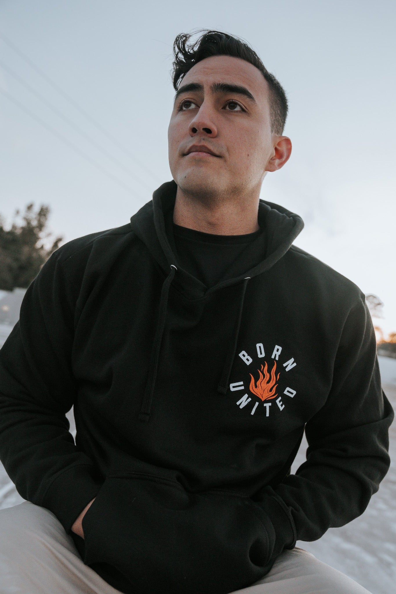 A person sits outdoors in daylight wearing a black Burning Bridges Hoodie from Born United Private Label, featuring Born United and a flame logo. With short dark hair, they gaze into the distance where trees stand silently in the background.