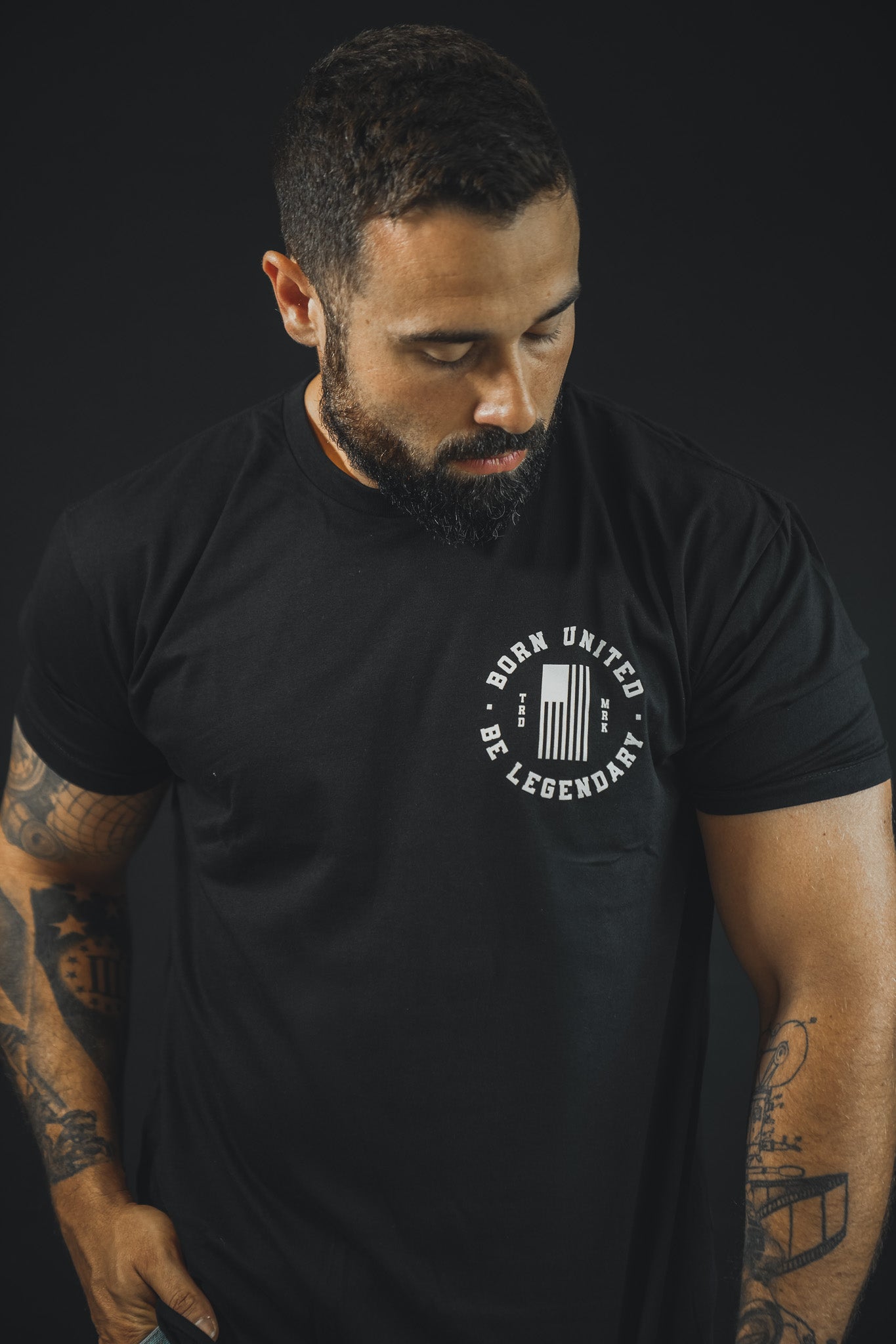 A bearded, tattooed man looks down in a dark setting, wearing the Born United Private Label "Legend - Washington" tee featuring BORN UNITED BE LEGENDARY and vertical bars—a bold tribute to American heritage.