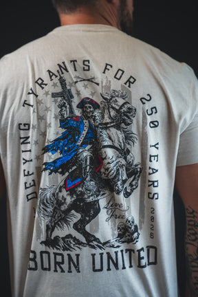 Someone wears the Born United Private Label 250th Anniversary Tee, featuring a colonial soldier on a rearing horse with a musket and text: “Defying Tyrants for 250 Years,” “Born United,” and “1776-2026,” celebrating American resilience.