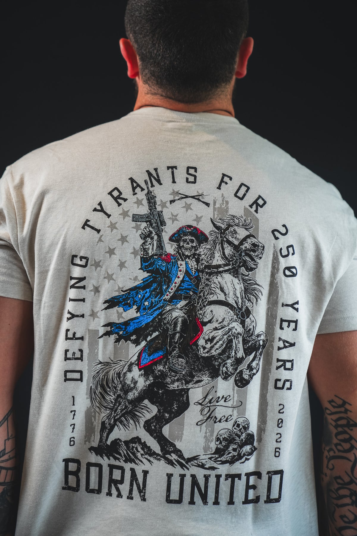 A man wears the Born United Private Label 250th Anniversary Tee, featuring a revolutionary soldier on horseback with "Defying tyrants for 250 years, 1776-2026, live free, born united." Celebrate American resilience in this special edition.
