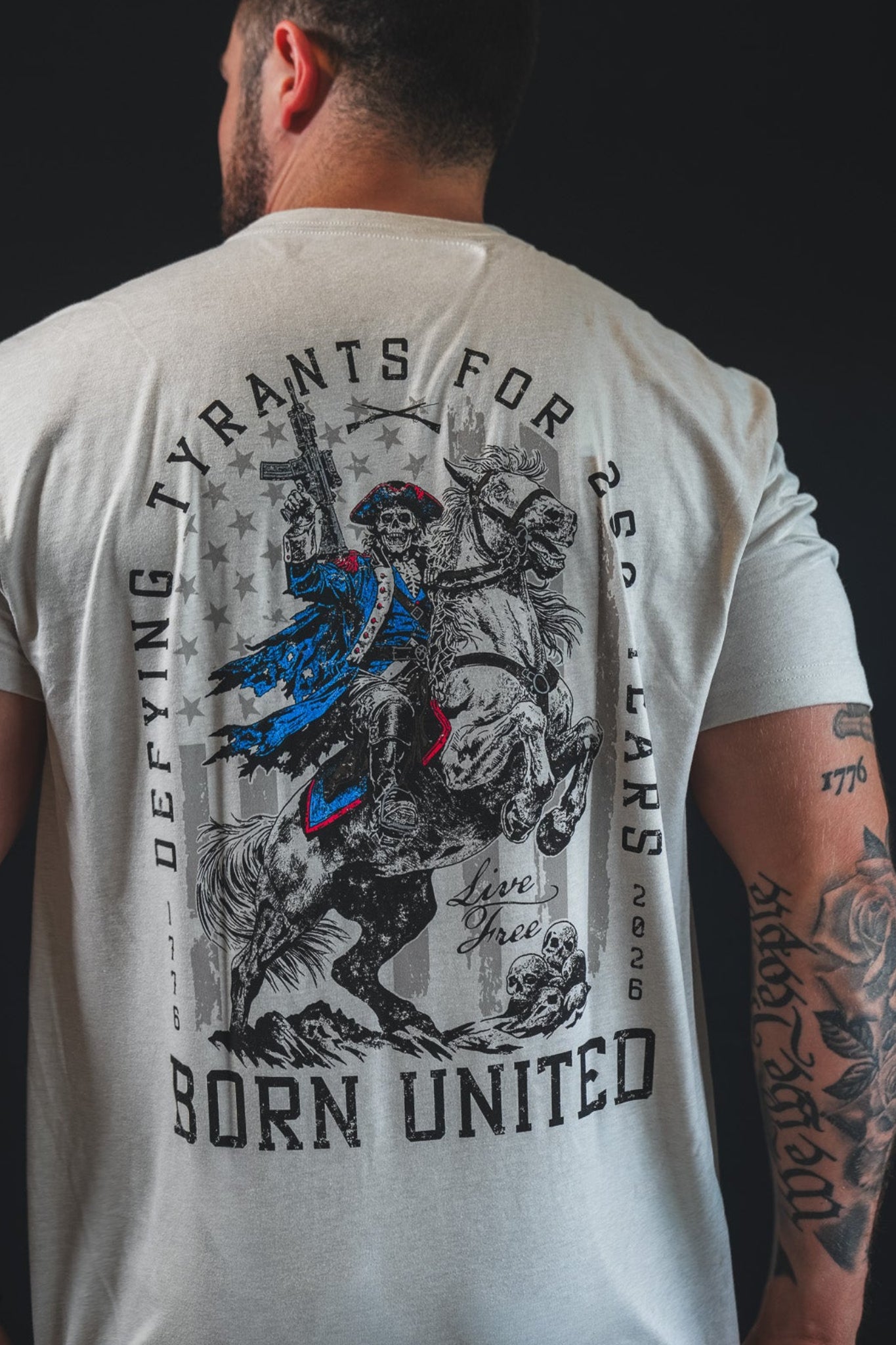 A tattooed man wears the Born United Private Label 250th Anniversary Tee, featuring a historical figure on horseback with a rifle and the slogan "Defying Tyrants for 250 Years," celebrating American resilience and independence.