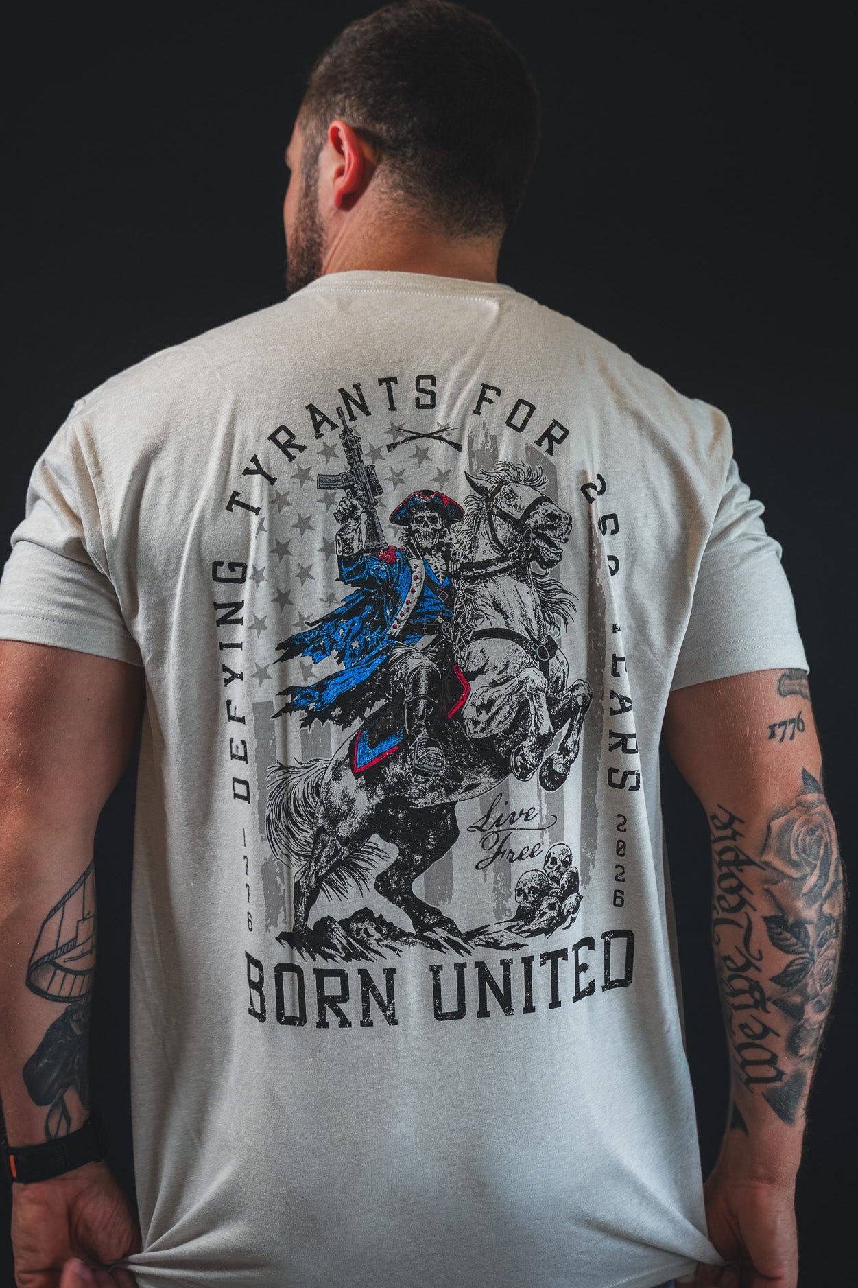 A tattooed man wears the 250th Anniversary Tee by Born United Private Label, featuring a rider, DEFYING TYRANTS FOR 246 YEARS, and BORN UNITED graphics.