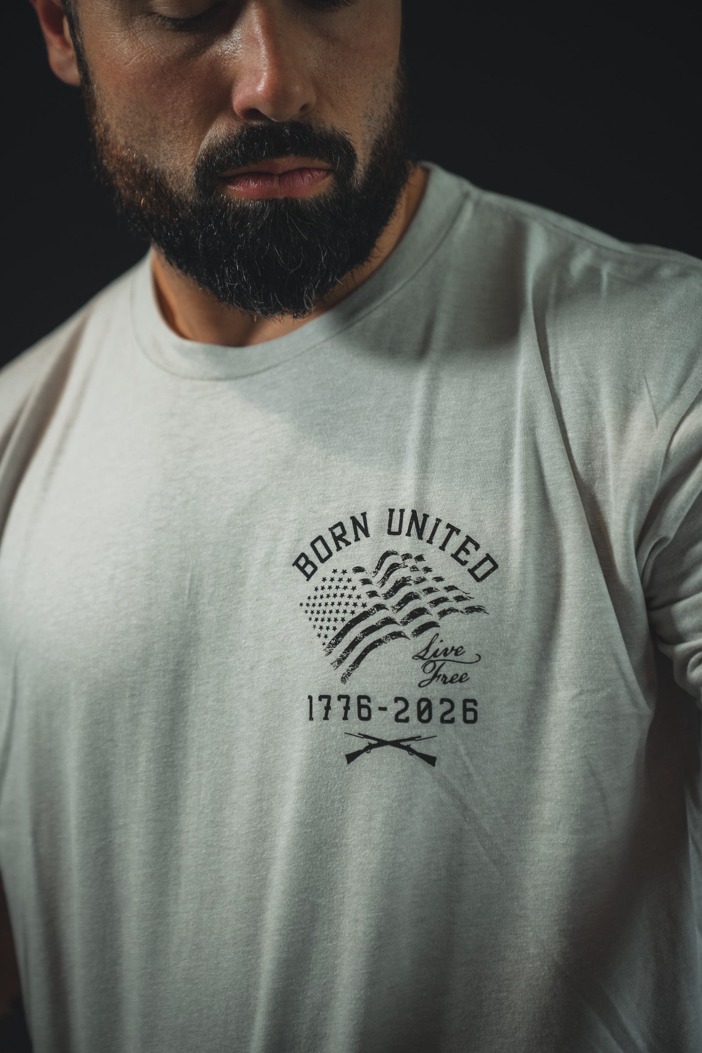 A bearded man wears the Born United Private Label 250th Anniversary Tee, a light t-shirt with a printed American flag and “BORN UNITED Live Free 1776-2026” on the chest, celebrating 250 years of American independence.