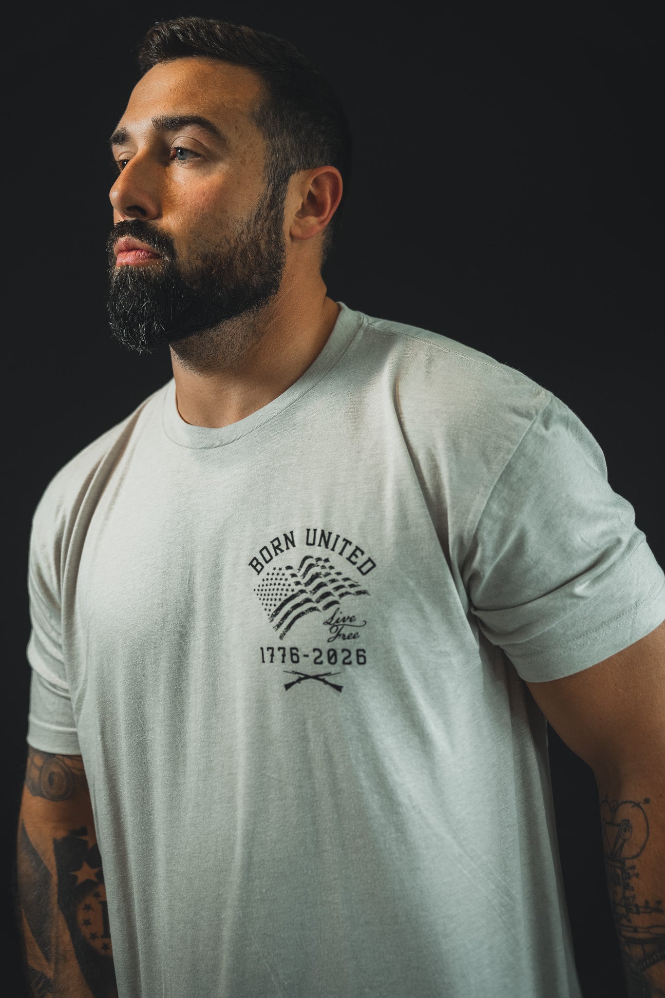 A bearded, dark-haired man with arm tattoos wears the Born United Private Label 250th Anniversary Tee, featuring "Born United 1776-2026" to celebrate American resilience. The background is dark and plain.
