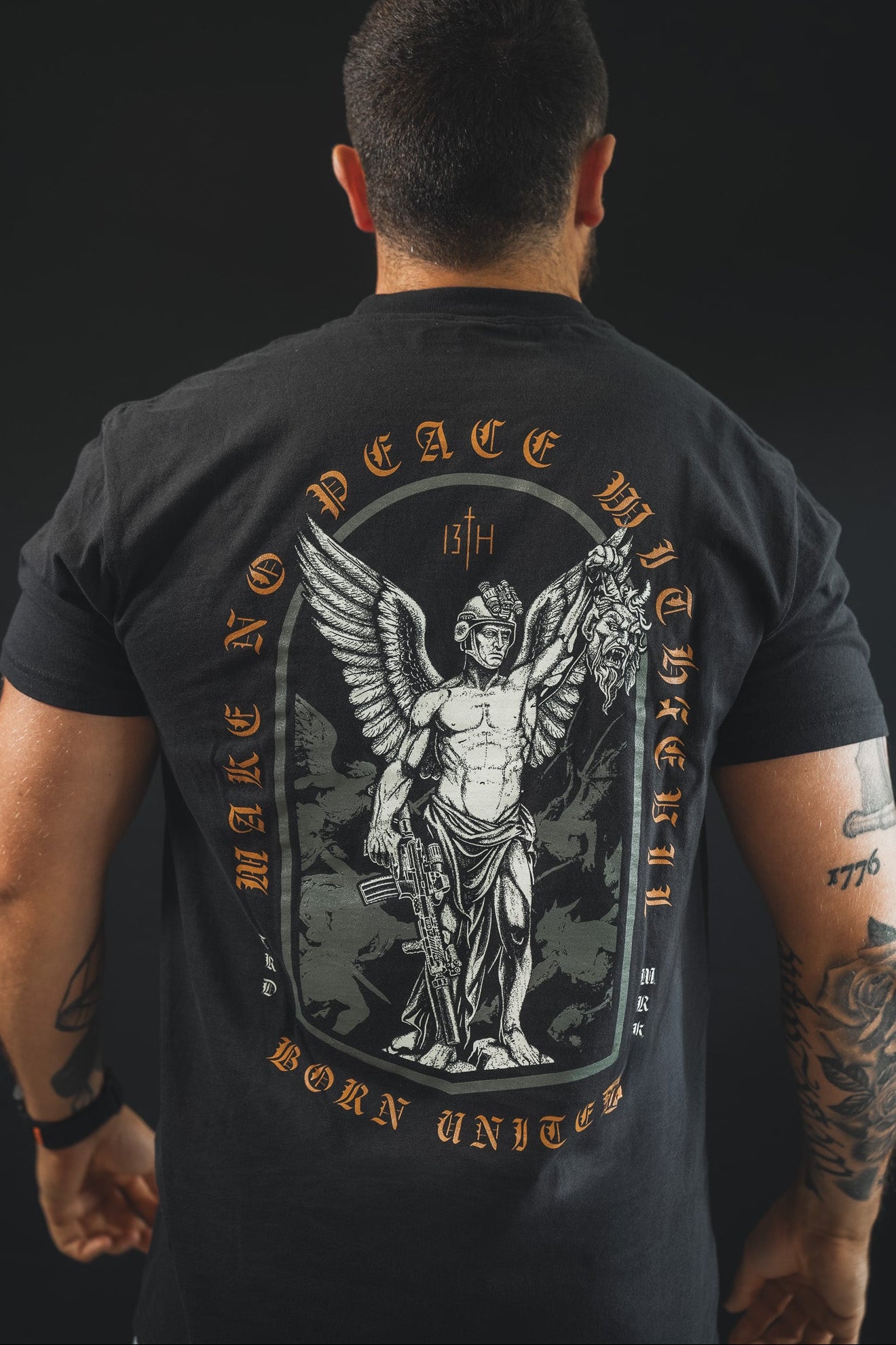 A man faces away wearing the Make No Peace Premium Tee by Born United Private Label—crafted from premium cotton, it features a winged warrior with sword and head, plus "MAKE NO PEACE WITH THE 13TH BORN UNITED" boldly printed on the back.