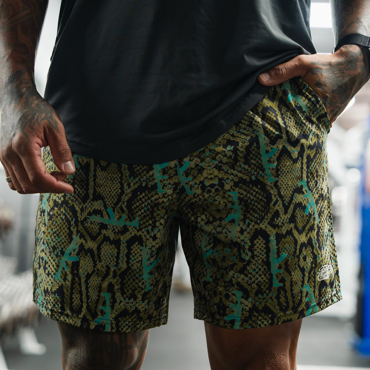 A person indoors lifts their black shirt to reveal Tango Charlie Apparel’s Men's Performance Training Shorts | 5.5"- Lethal Venom, featuring a striking green, yellow, and black snakeskin pattern.