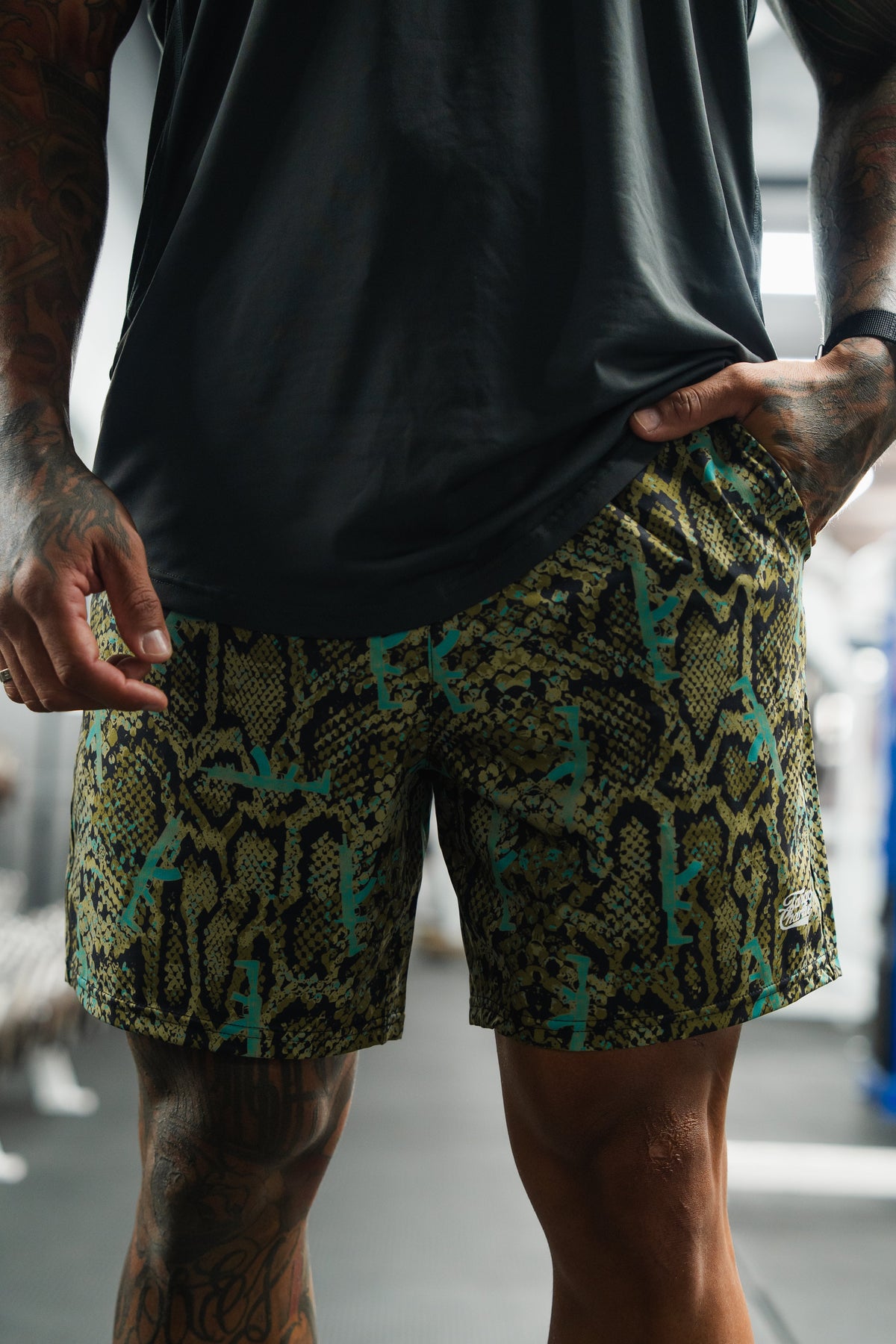 A person with tattooed arms and legs stands in the gym, hands at their sides, wearing a dark shirt and Tango Charlie Apparel Men's Performance Training Shorts 5.5" in the Lethal Venom colorway.