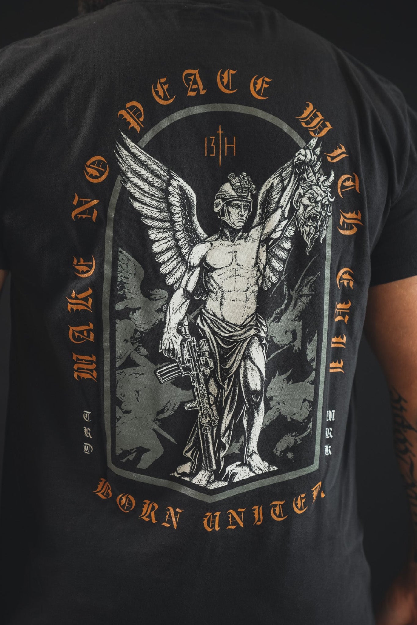 Someone wears the 13th Make No Peace Premium Tee, a bold statement shirt with a winged warrior, rifle, and severed head graphic. Bright orange text declares MAKE NO PEACE WITH EVIL and BORN UNITED on soft premium cotton.