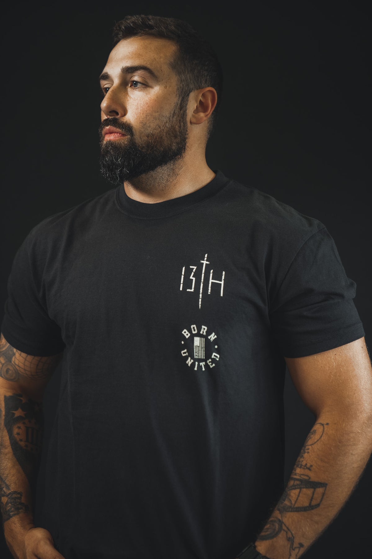 A bearded, tattooed man wears the 13th Make No Peace Premium Tee against a dark background, with BORN UNITED and 13TH printed on the chest as he looks to the side.