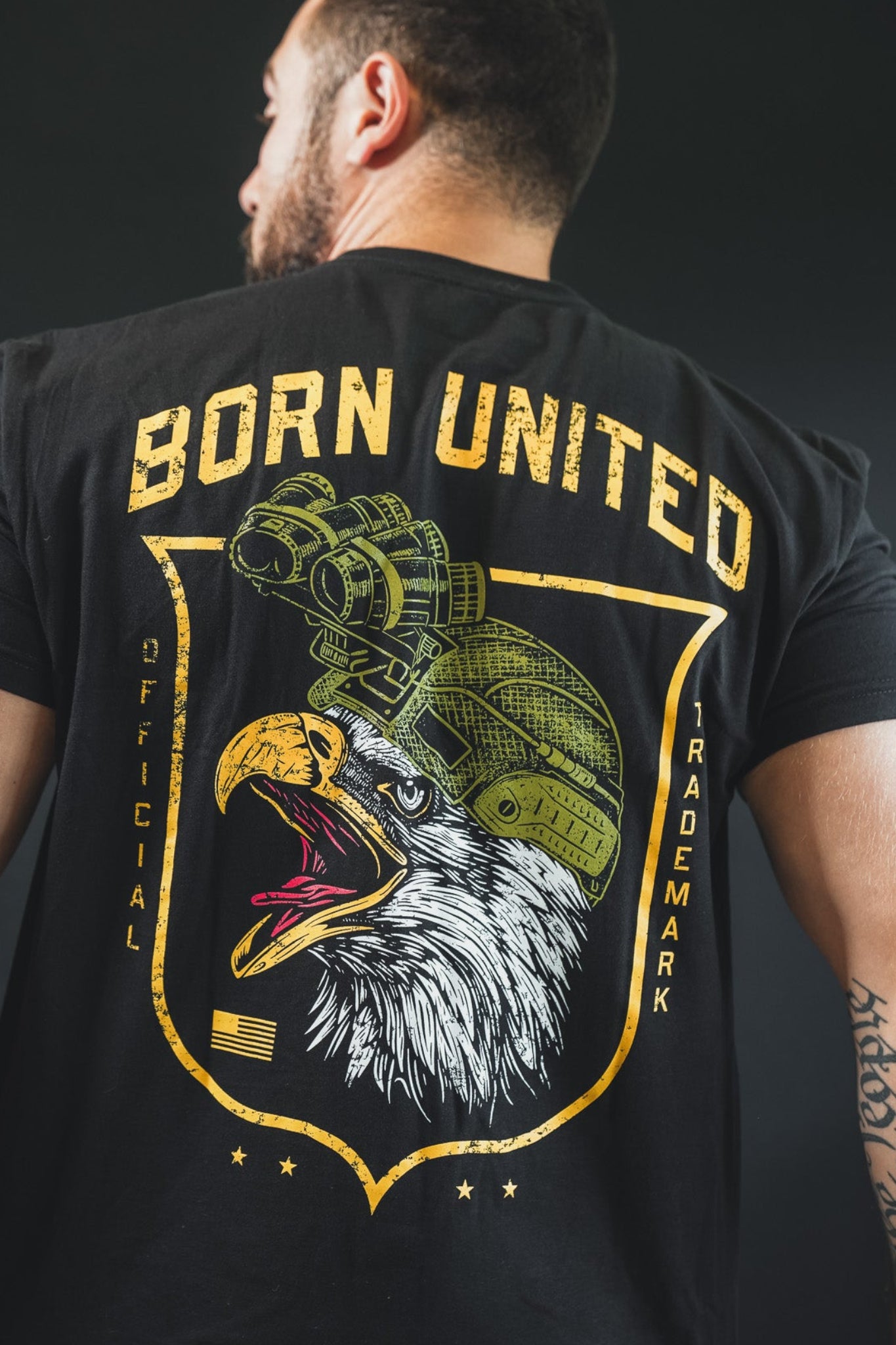 A tattooed man wears The Eagle's Scream tee from Born United Private Label, featuring a bold bald eagle in night vision goggles and "BORN UNITED" in yellow above, showcasing American pride.