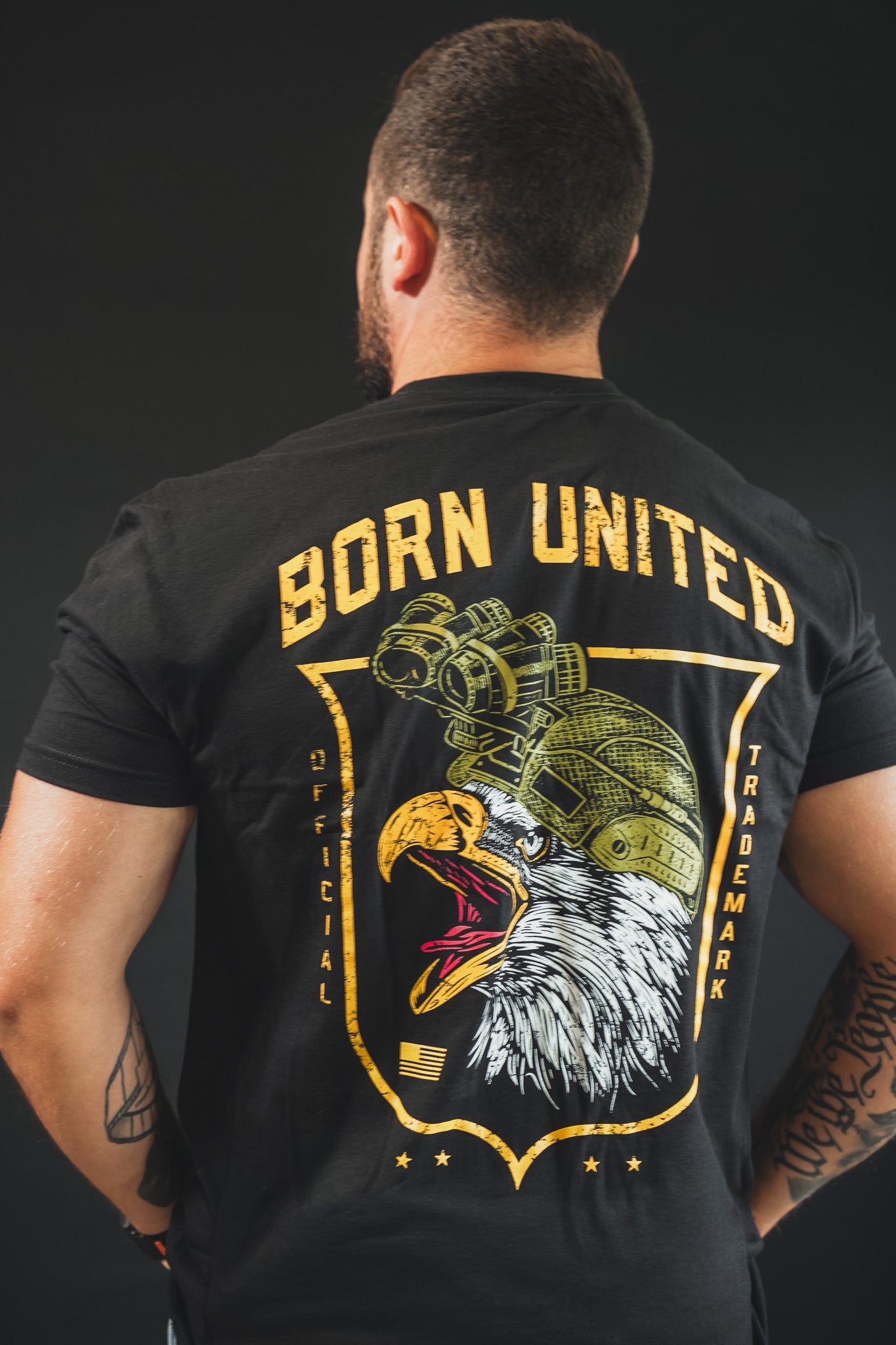 A tattooed man stands facing away, wearing The Eagle’s Scream tee by Born United Private Label—a black shirt with a bold bald eagle in a military helmet and "BORN UNITED" in yellow above, showing his American pride.
