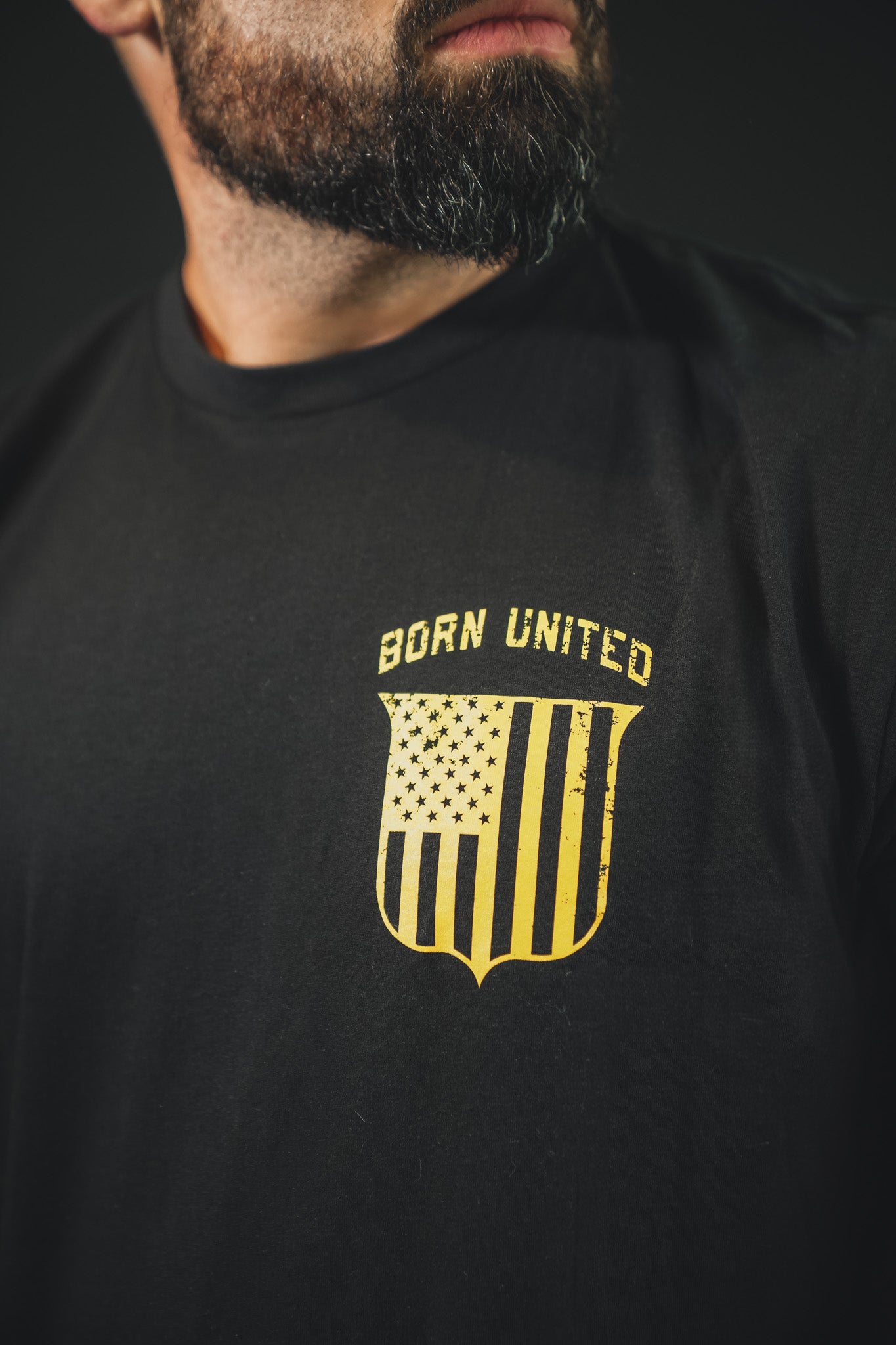 A bearded man wears "The Eagle's Scream"—a black tee by Born United Private Label, featuring a yellow shield with vertical stripes, stars, and the words BORN UNITED above it.