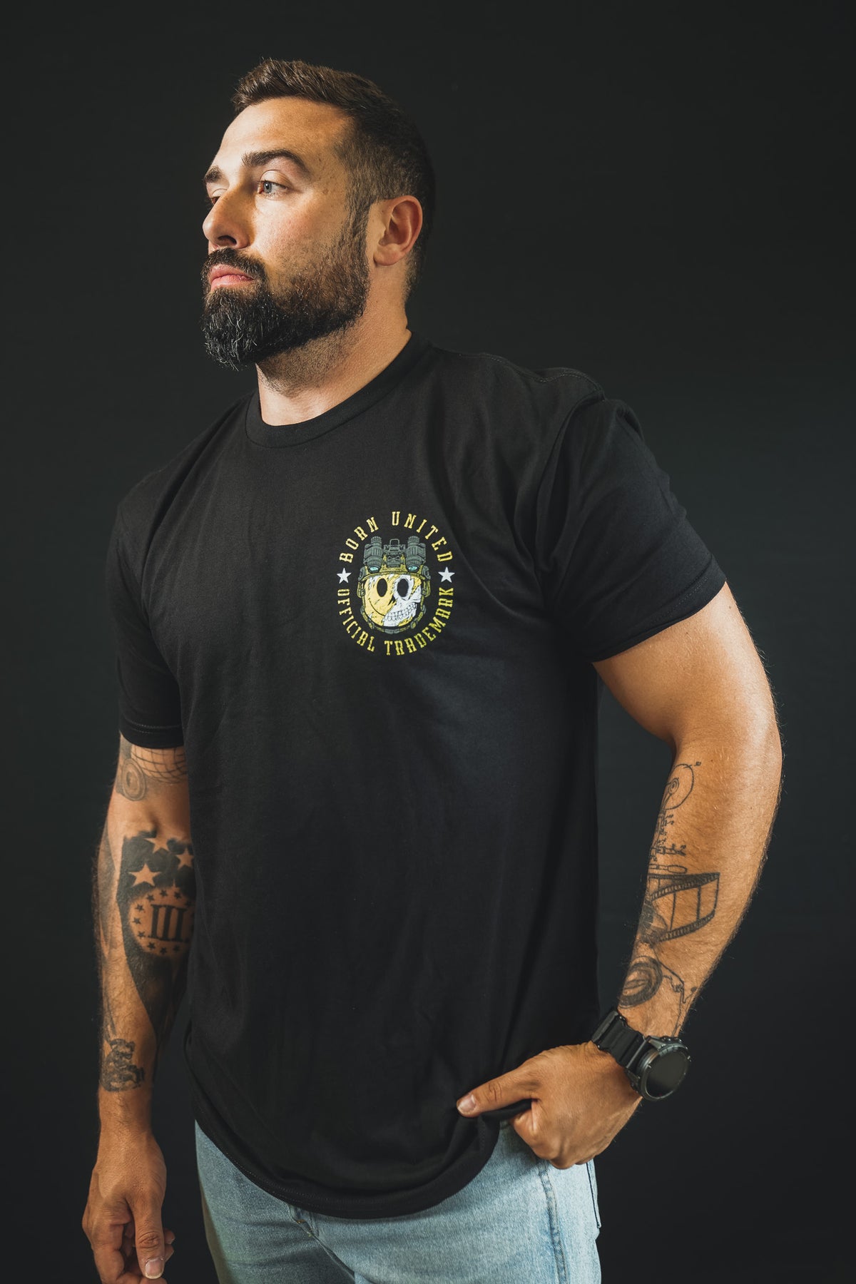 A bearded, tattooed man in a black Born United Private Label "Smile Back" tee and light jeans stands against a black background, gazing confidently to the side with one hand in his pocket.