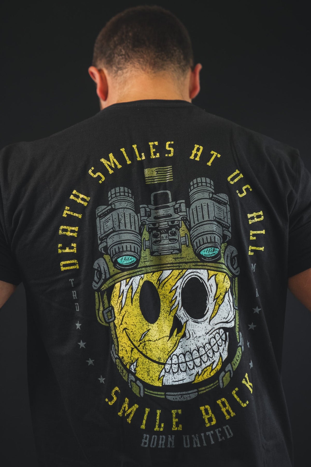 A man faces away, wearing the Born United Private Label "Smile Back" t-shirt featuring a skull half-covered by a yellow smiley face with night vision goggles and the text: Death smiles at us all. Smile back. Apparel for the bold.