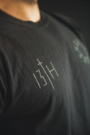 A close-up shows someone wearing the 13th Victorious Dawn Premium Tee, featuring white symbols and text on black fabric. The design stands out against a dark, blurred background, evoking spiritual strength.