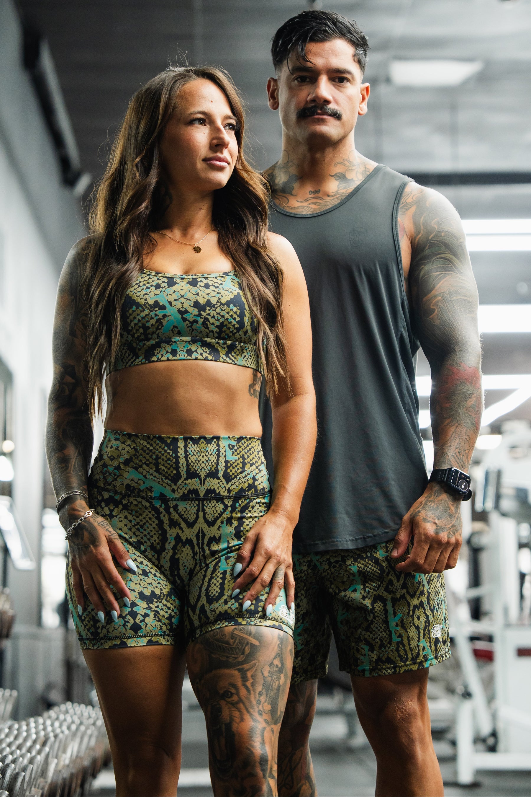 A woman and man pose confidently in a gym, wearing matching green and black Tango Charlie Apparel outfits. He showcases the Men's Performance Training Shorts | 5.5" – Lethal Venom, highlighting sporty style and fitness readiness.