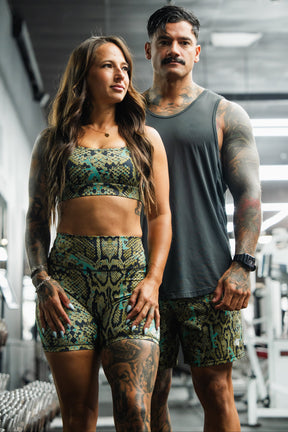 A woman and man pose confidently in a gym, wearing matching green and black Tango Charlie Apparel outfits. He showcases the Men's Performance Training Shorts | 5.5" – Lethal Venom, highlighting sporty style and fitness readiness.