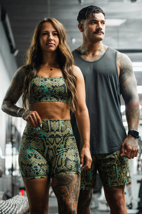 A fit woman and man stand side by side in a gym. She wears the Tango Charlie Apparel Straight Strap Sports Bra | Lethal Venom; he wears a gray tank and shorts. Both have tattoos and serious expressions.