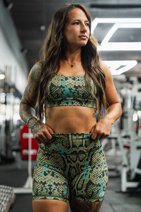 A woman stands confidently in a gym wearing the Tango Charlie Apparel Straight Strap Sports Bra | Lethal Venom, paired with matching shorts. Exercise equipment and weights are visible in the background as she looks to the side, hands on hips.